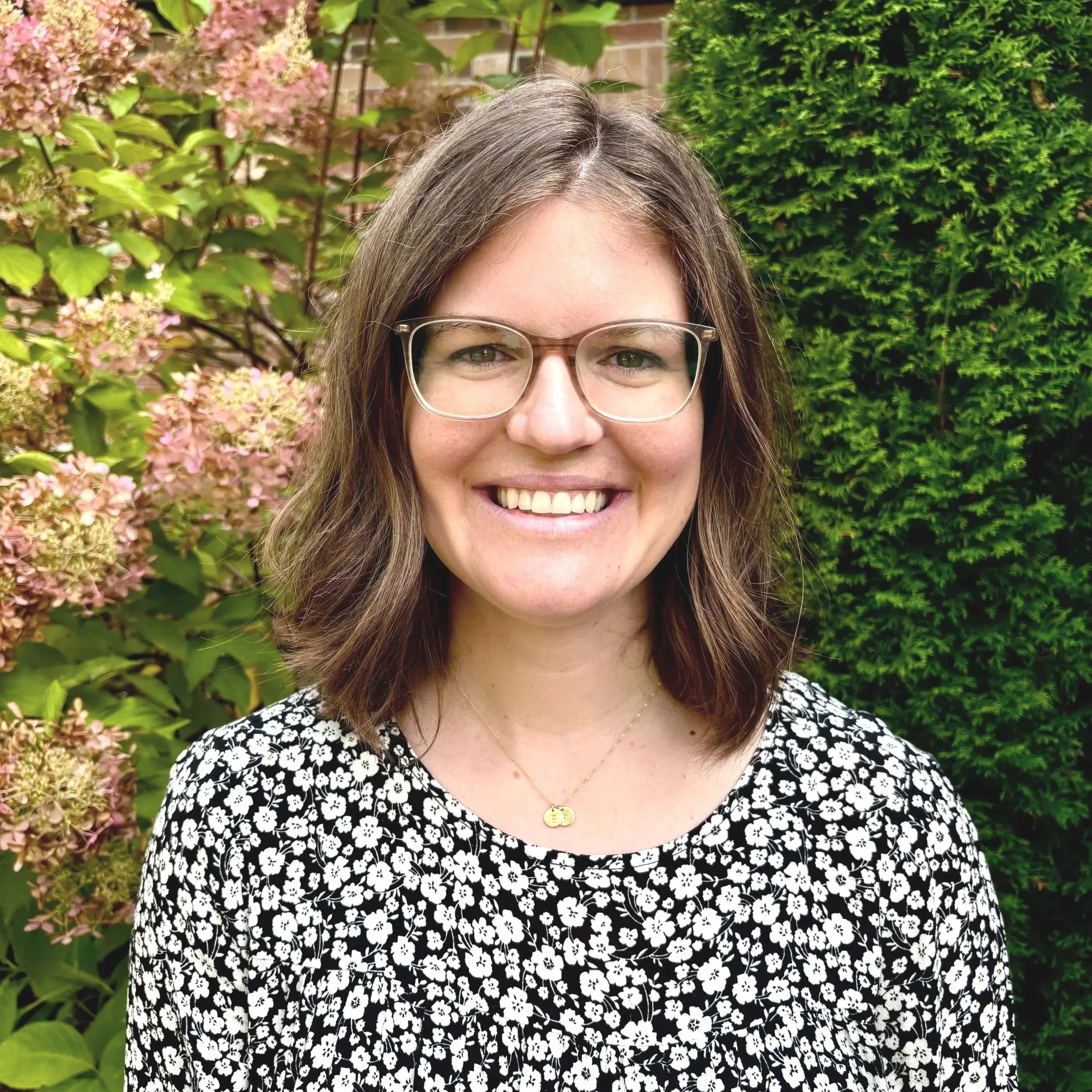 Smiling woman wearing glasses and a black-and-white floral top, standing outdoors in a garden.