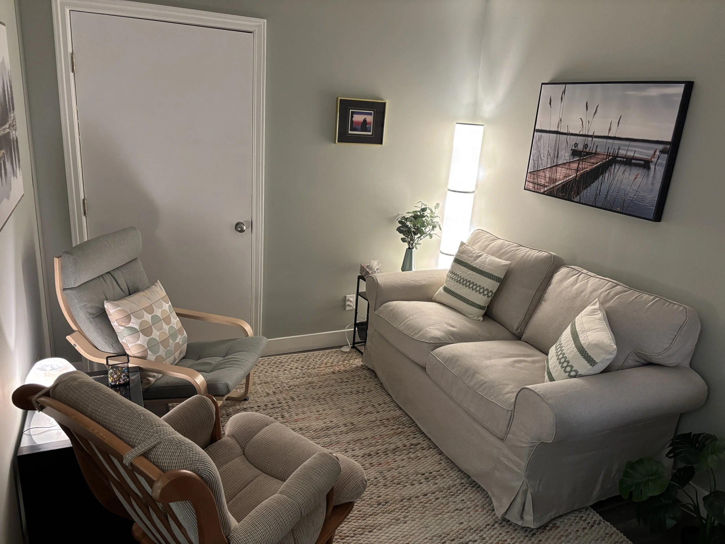 Cozy living room with a gray sofa, two wooden lounge chairs, patterned cushions, floor lamp, potted plants, area rug, and framed lakeside artwork.
