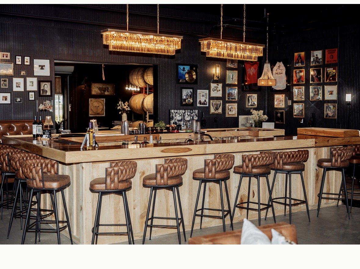 Interior view of a modern bar with a large wooden counter, leather bar stools, and a black wall decorated with framed pictures, artwork, and vintage decorations. Hanging lights add warm lighting to the space.