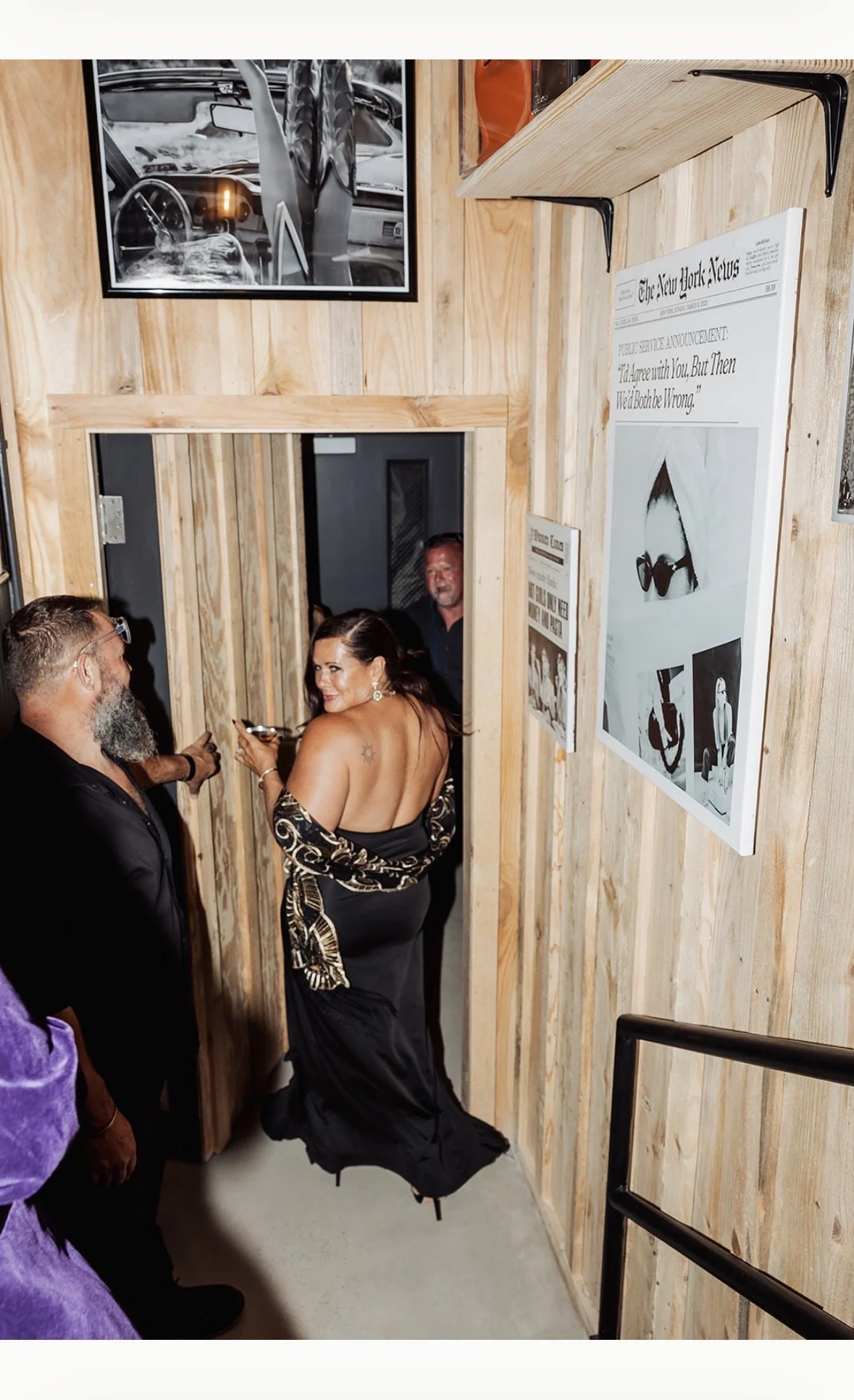 People in formal attire standing in a wooden hallway with framed pictures and a newspaper mounted on the wall.