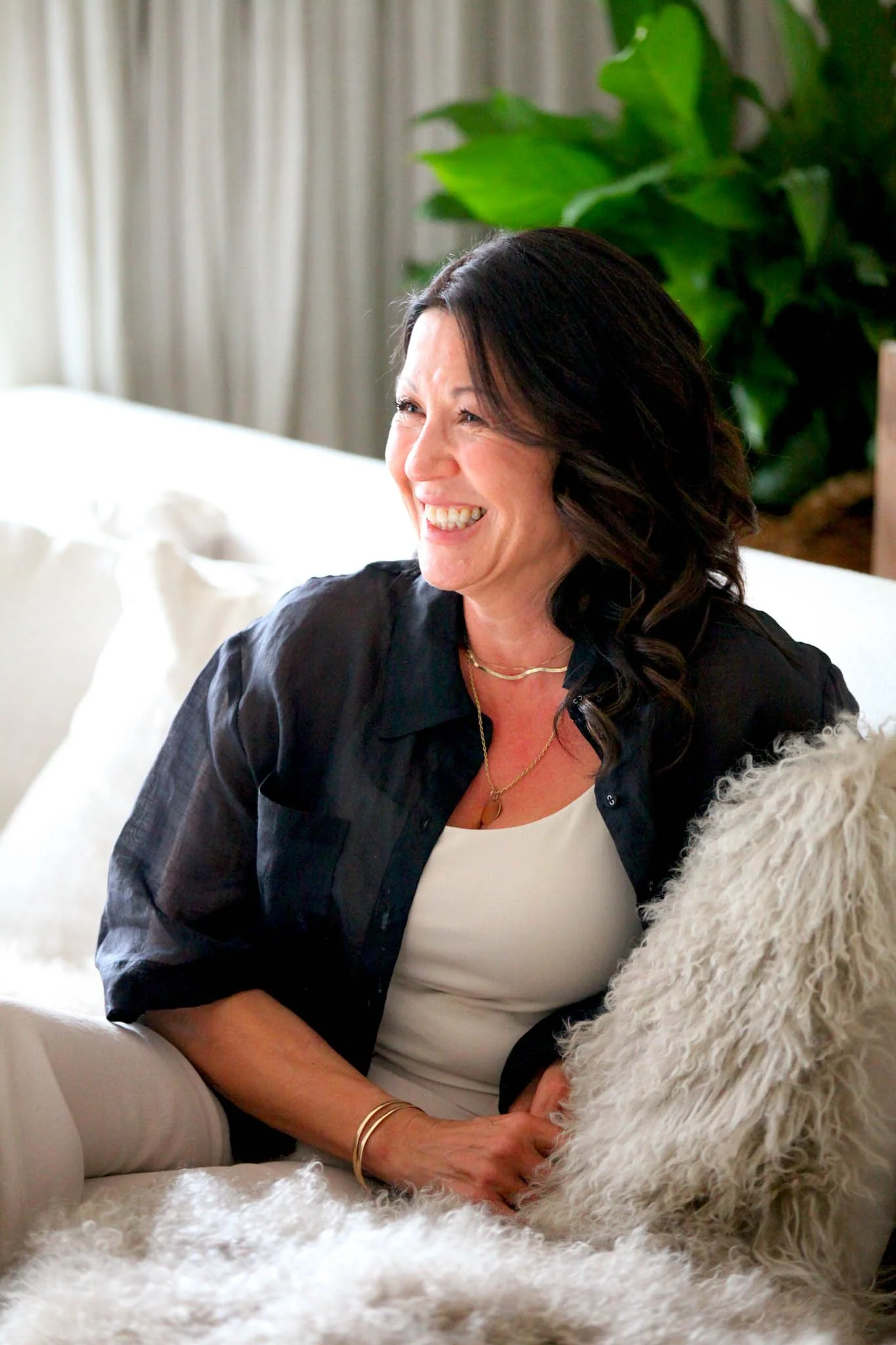 A woman with dark hair smiling and sitting on a white couch, with a large plant in the background and a furry white dog laying nearby.