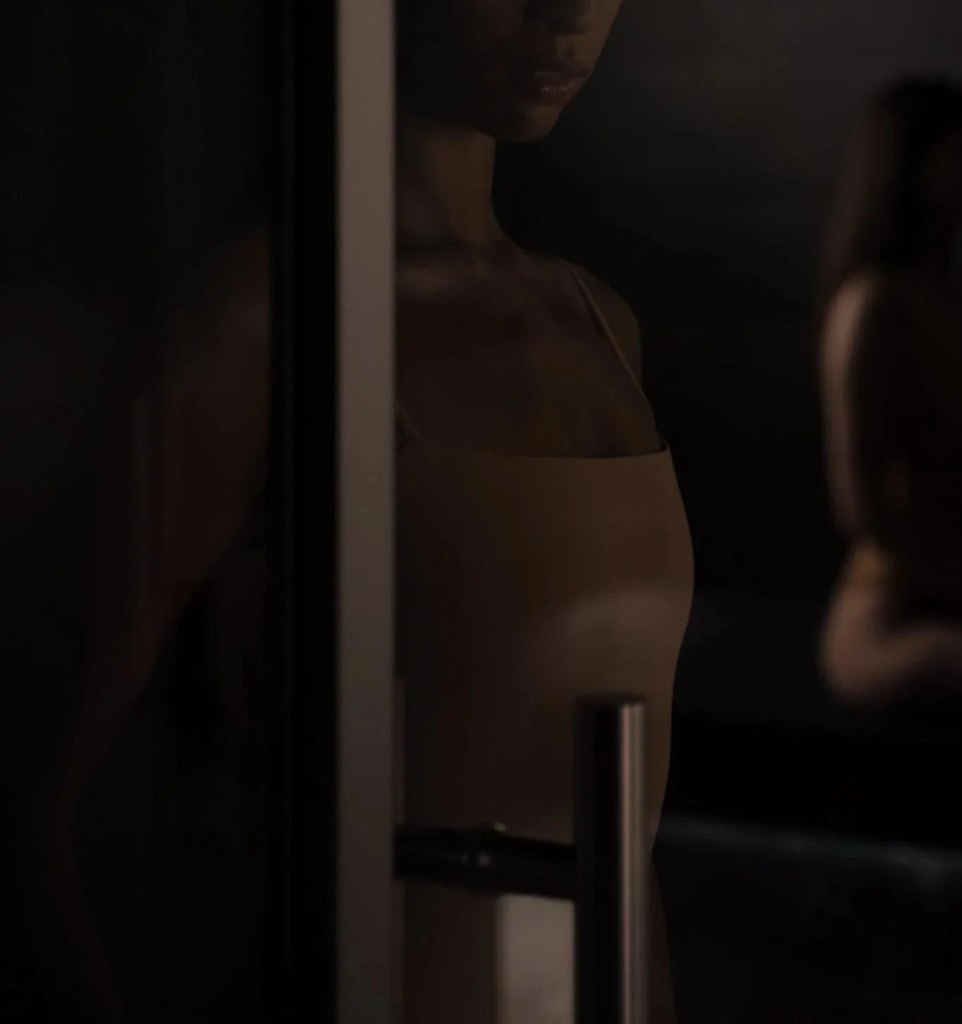 A dark photo of a woman with dark hair, wearing a beige top, partially visible with a focus on her profile and shoulder, with blurred objects and shadowed figures around her.