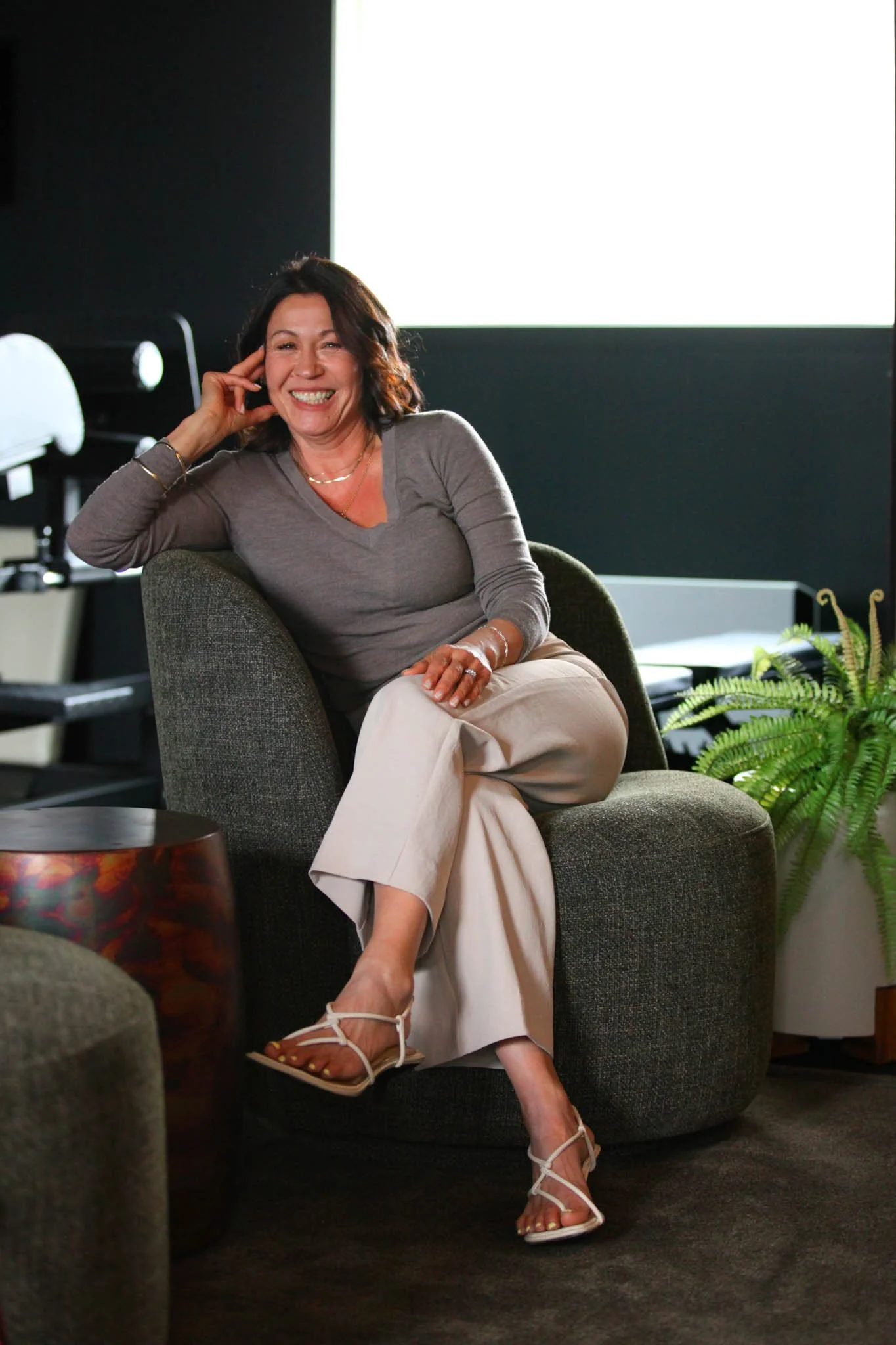 A woman sitting on a dark gray armchair, smiling and resting her head on her hand. She has shoulder-length dark hair and is wearing a gray long-sleeve top, beige pants, and white heeled sandals. There is a potted fern plant on her right and a white screen or window in the background.