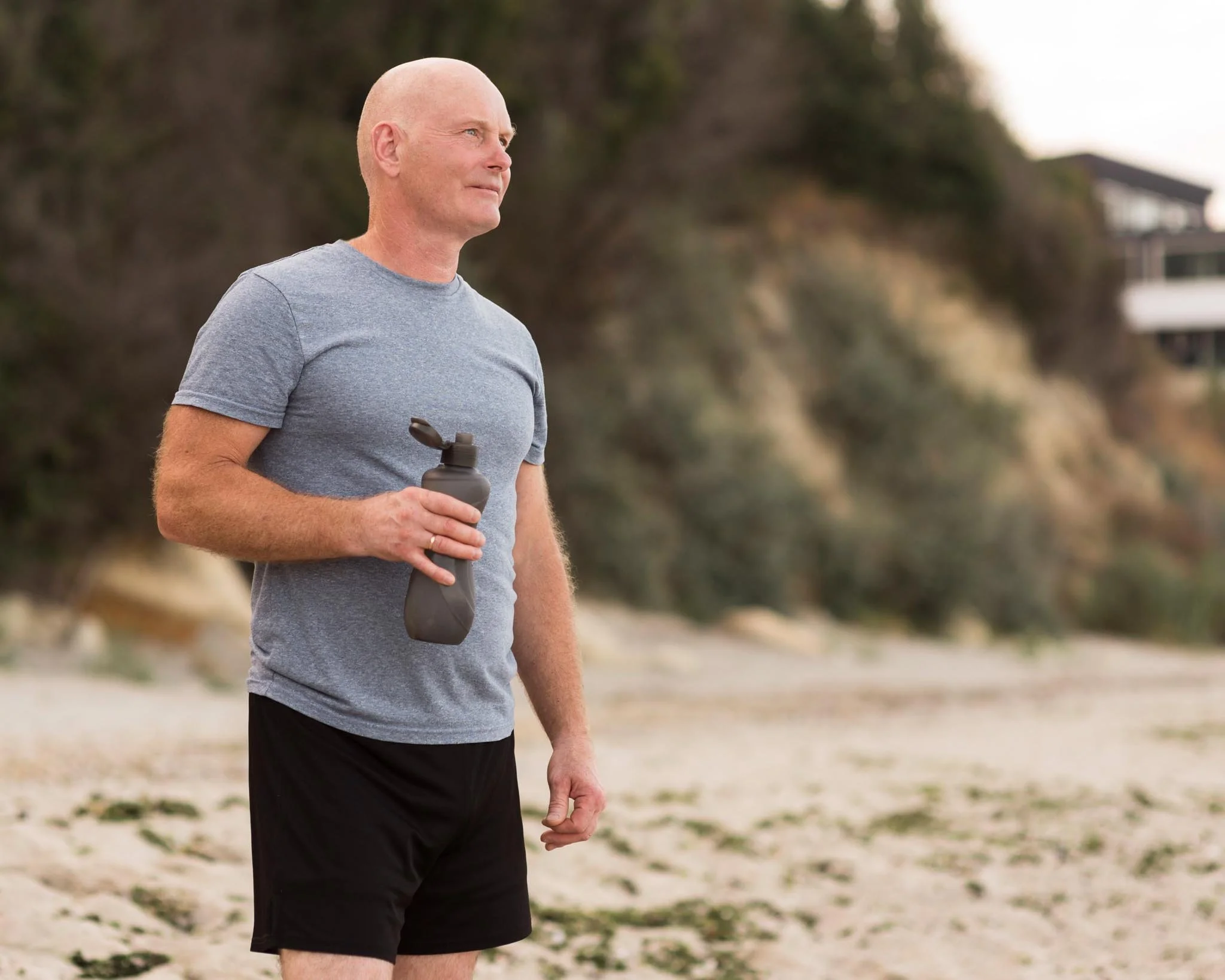A middle-aged man with a shaved head, wearing a gray T-shirt and black shorts, walking outdoors on a sandy beach, holding a water bottle in his right hand, with blurry trees and a building in the background.
