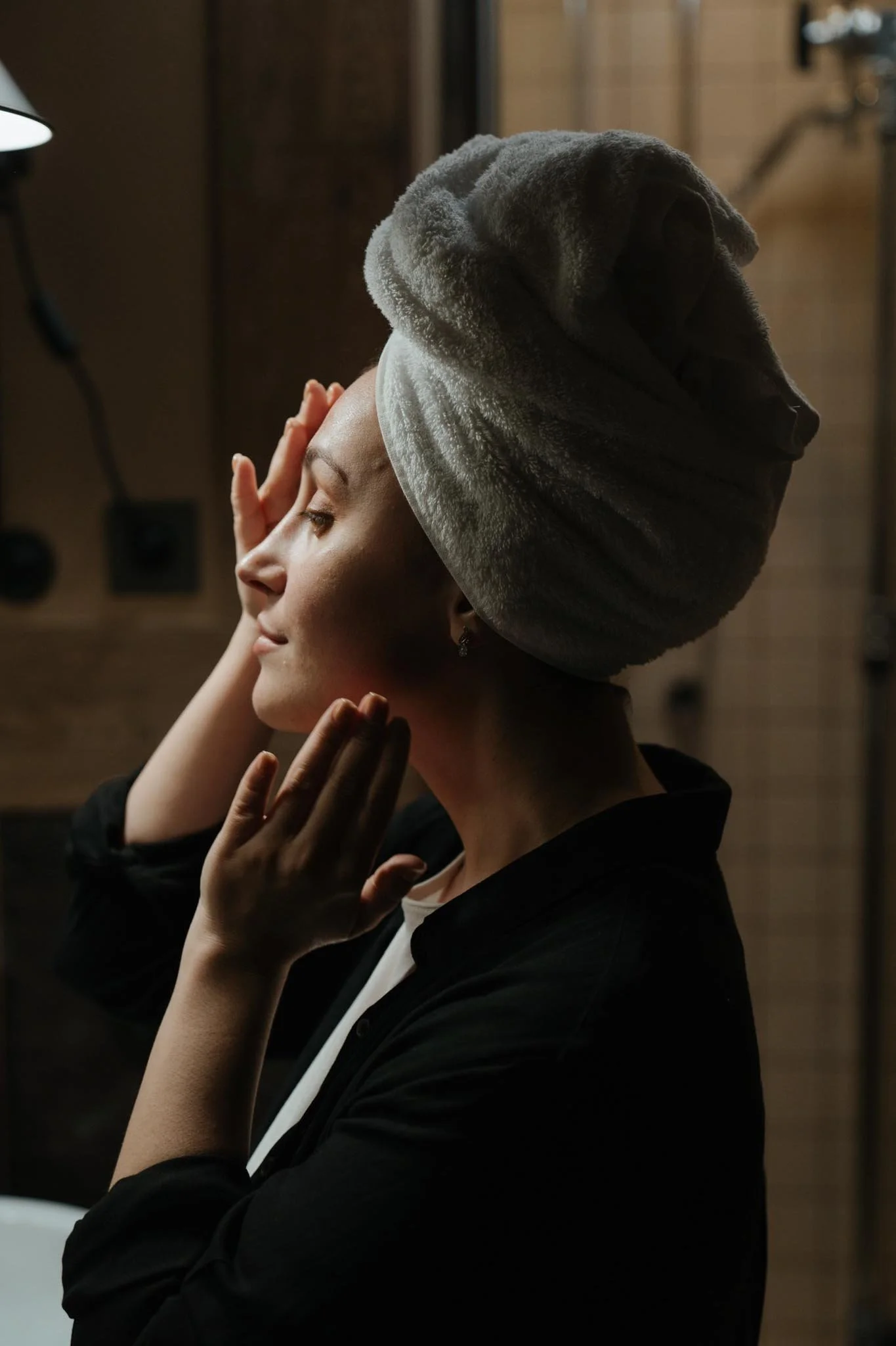 A woman with her eyes closed, gently touching her face with her hands, wearing a towel wrapped around her head.