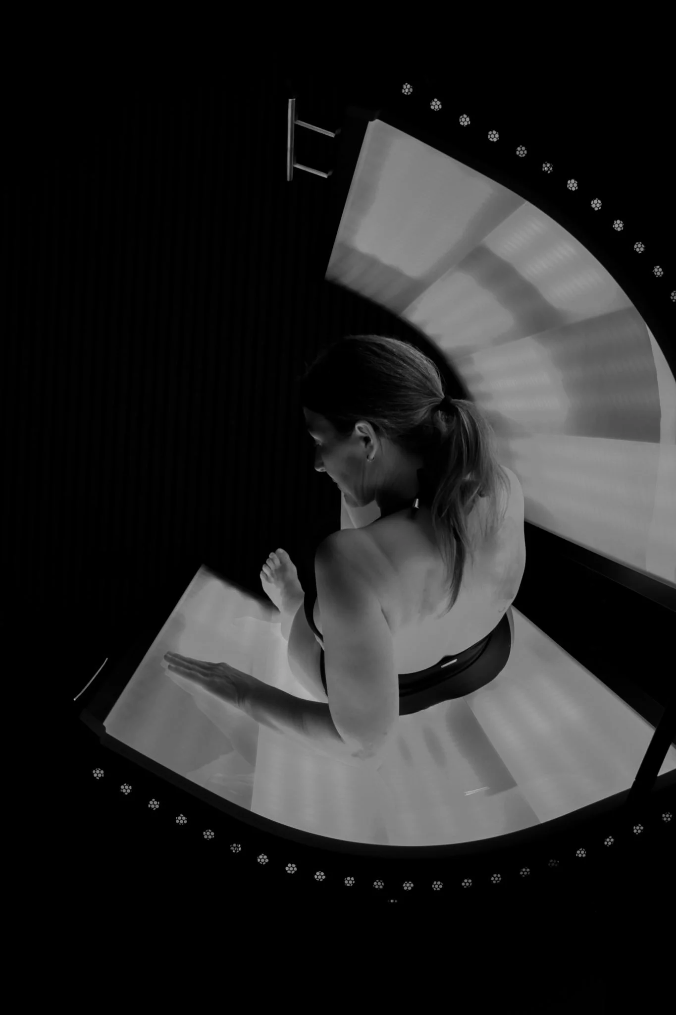 Black and white photo of a woman practicing yoga in a studio with a large circular window behind her, reflecting light onto her back while she sits on a yoga mat with one arm extended.
