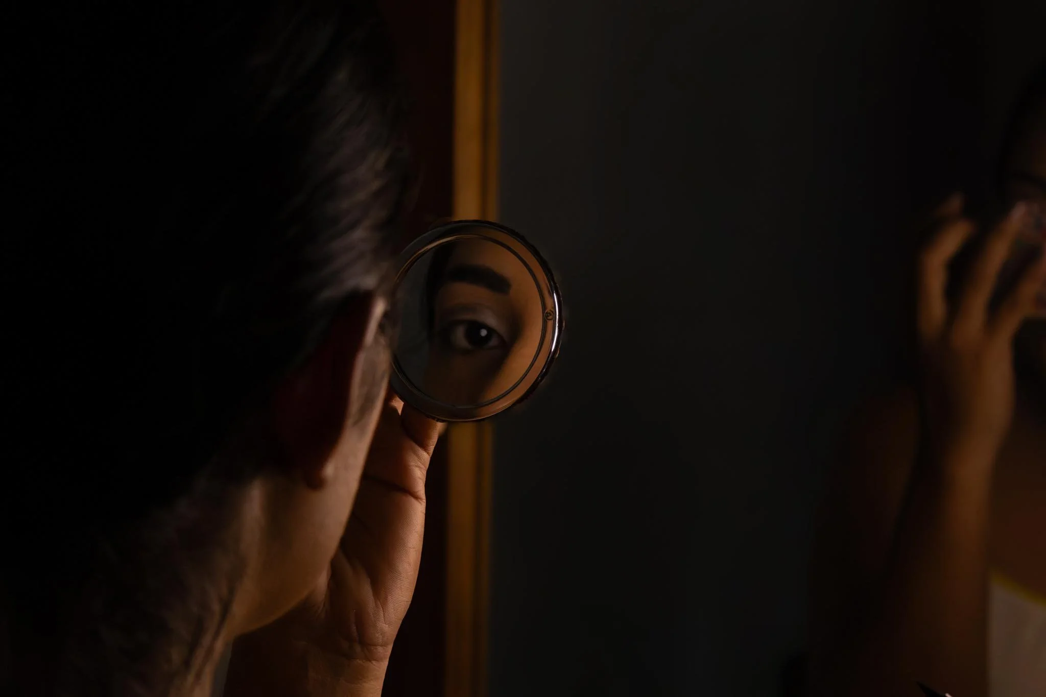 A person holding a small mirror reflecting their eye, with a dark background and part of a figure’s head visible.