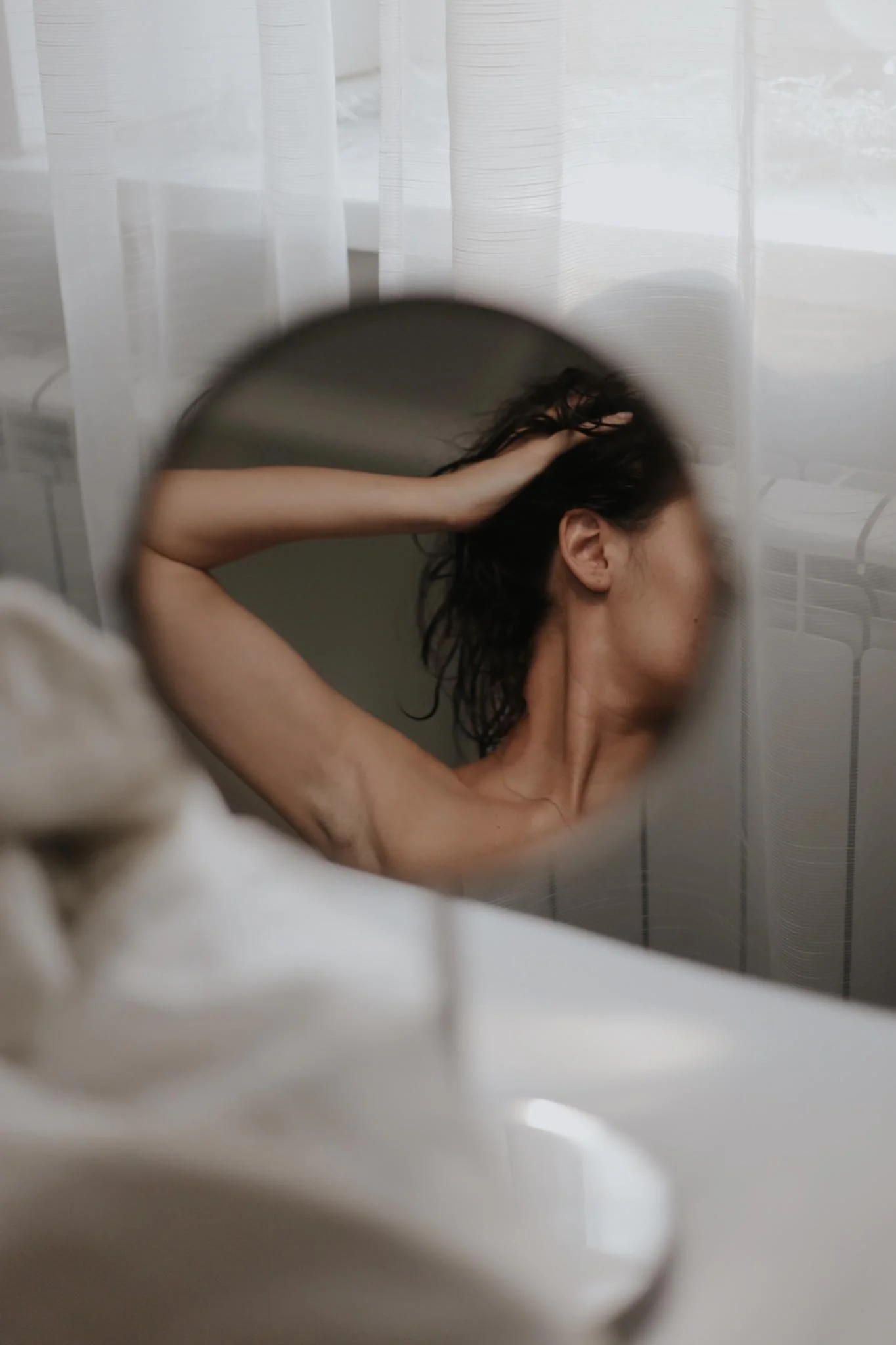 A person with wet, dark hair, seen through a round mirror, is touching their head with one hand and appears to be in a bathroom with white curtains.