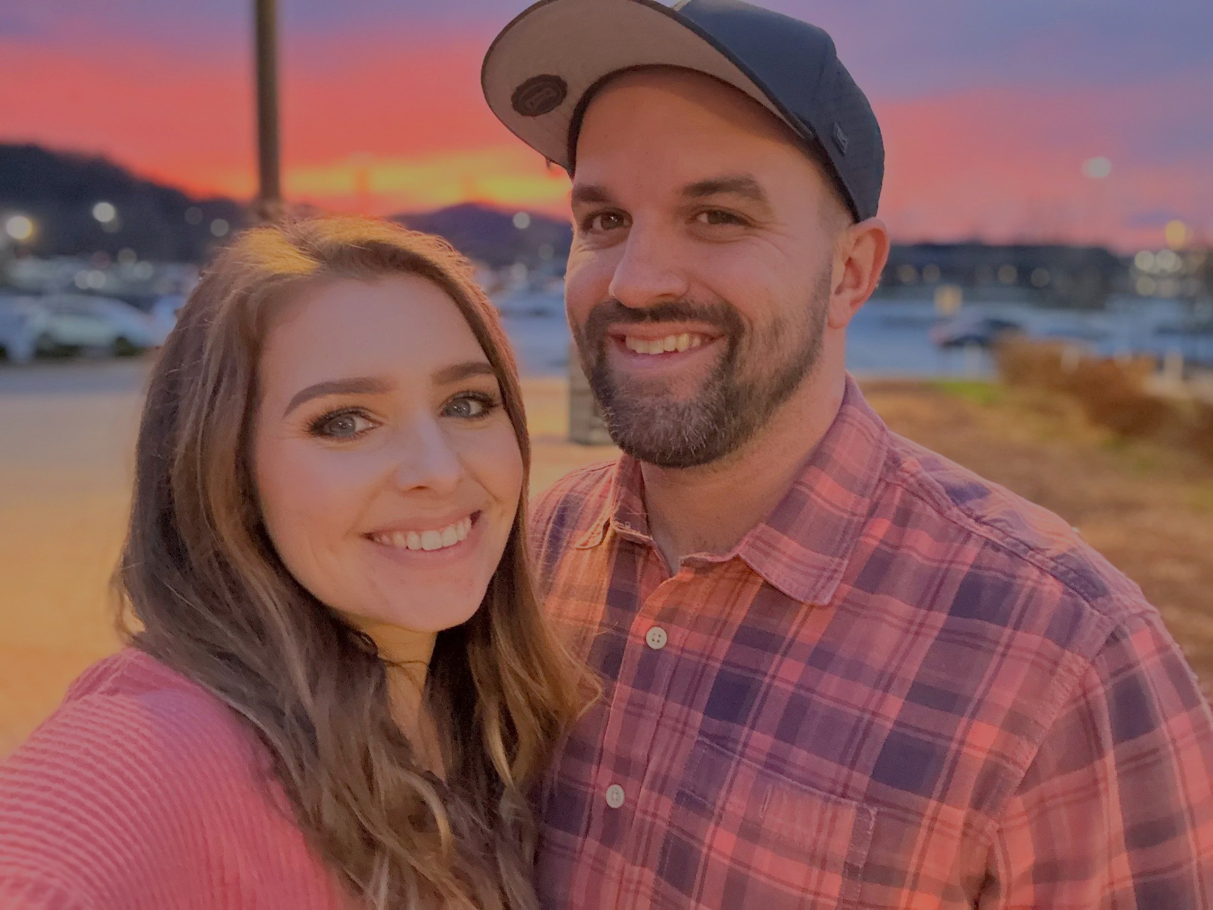 A smiling woman and a smiling man taking a selfie outdoors during sunset, with boats and water in the background.