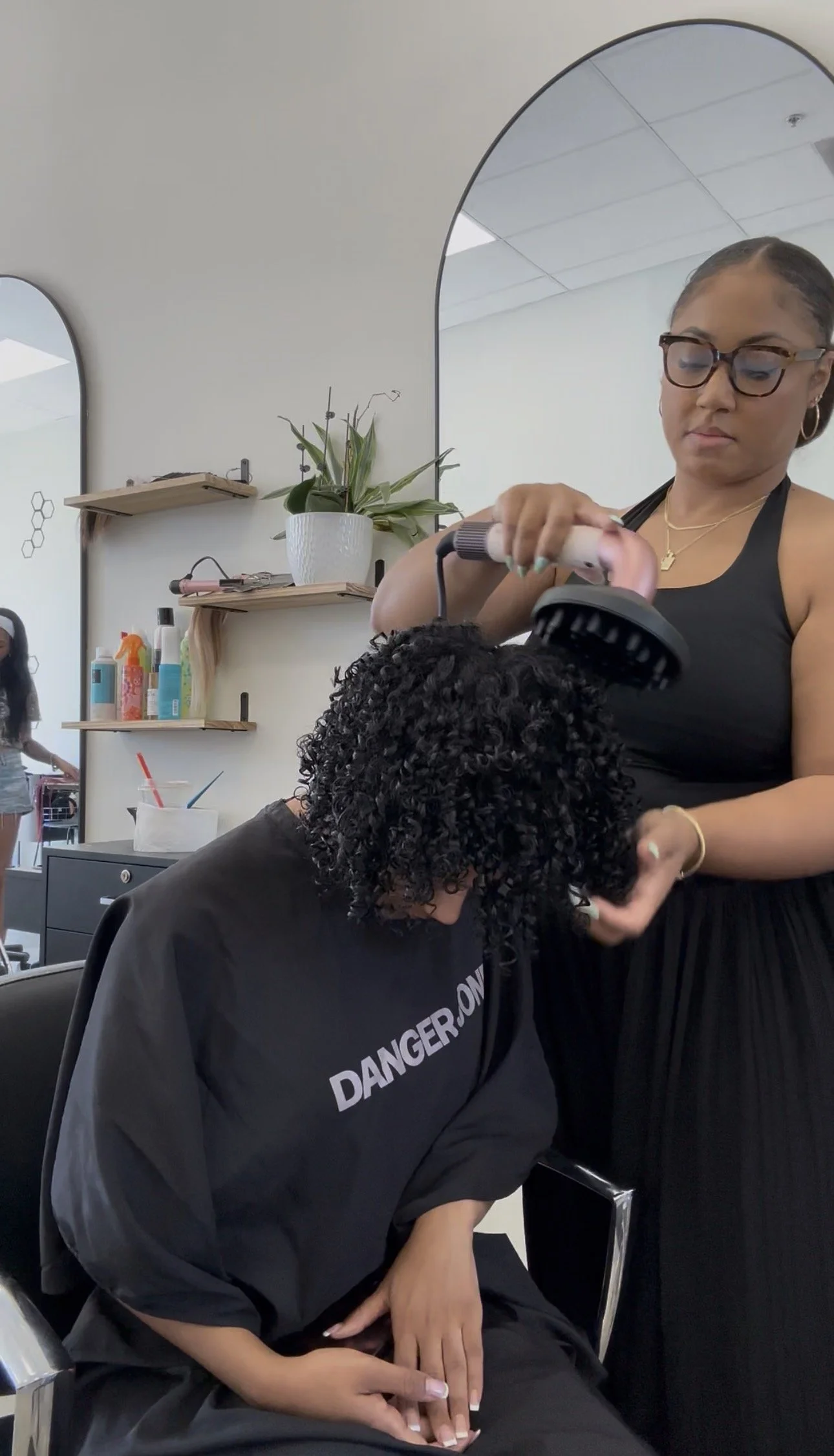 A hairstylist is blow-drying a woman's curly hair in a salon. The stylist is wearing glasses and a black tank top, with a mirror and salon products visible in the background.