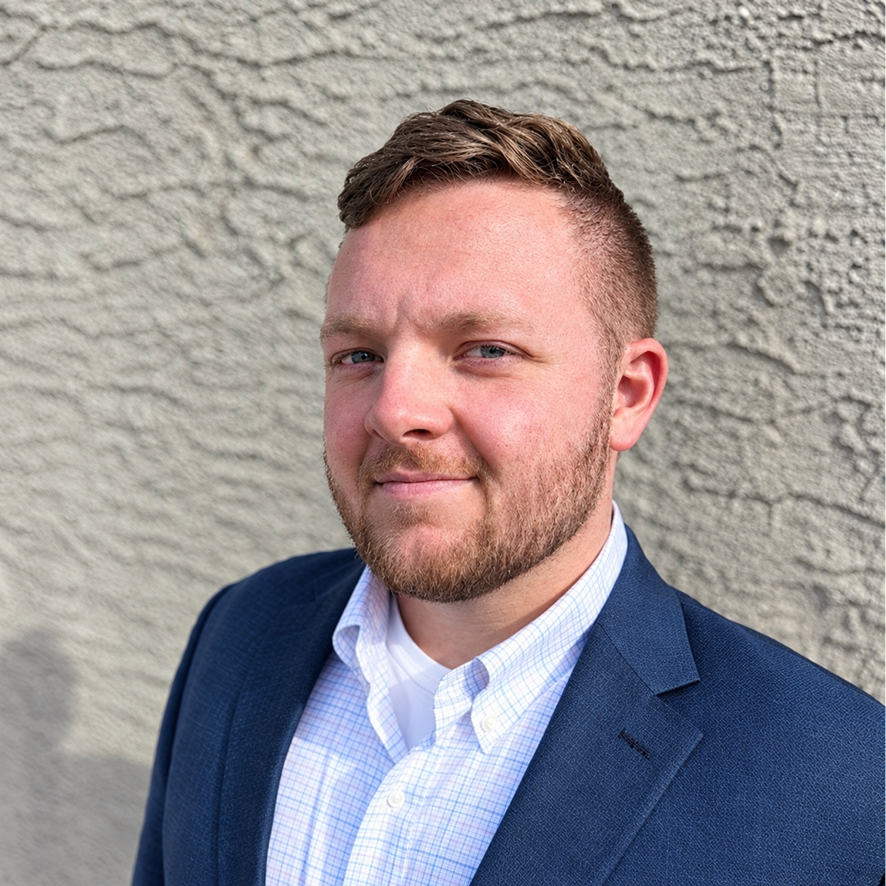Max Williams, founder of Tailwind Unmanned. Headshot of a young man with short brown hair, light facial hair, wearing a navy blazer and a light blue checkered shirt, standing in front of a textured gray wall.