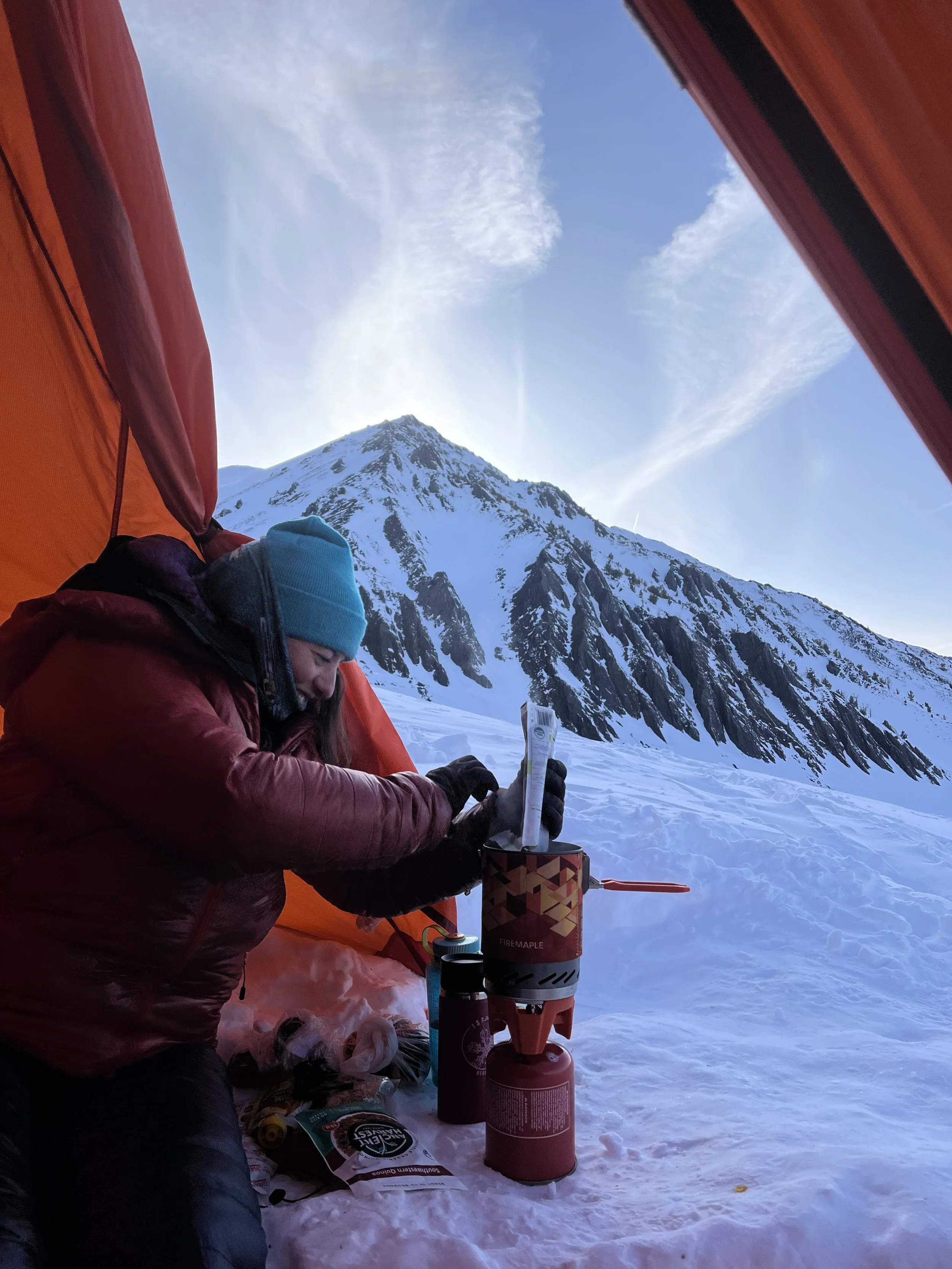 Person in a winter jacket and blue beanie sitting inside a tent, with snow and a mountain outside, preparing a small camping stove with a propane tank.