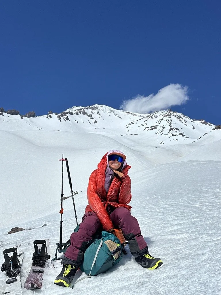 Person in winter gear sitting on snow with mountains in the background, ski equipment on the snow, wearing sunglasses, red jacket, purple pants, and smiling.