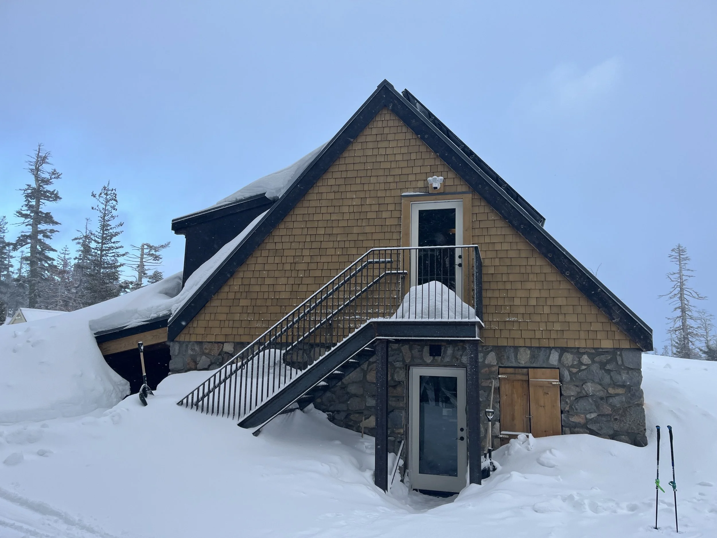 A house in a snowy landscape with brown shingle siding and black trim. A staircase leads up to a door on the second floor. Snow covers the roof and ground, with trees in the background and a cloudy sky.