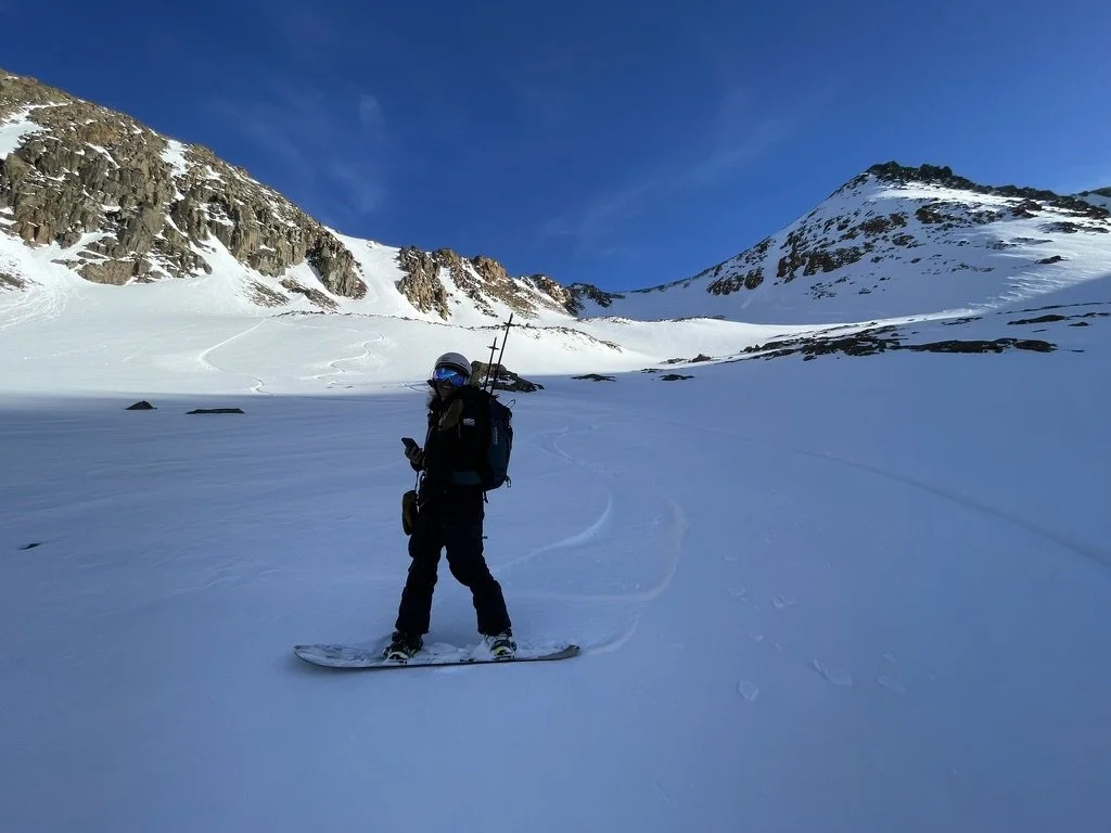 A person in snow gear standing on a snowboard in a snowy mountain landscape with rugged peaks and a clear blue sky.