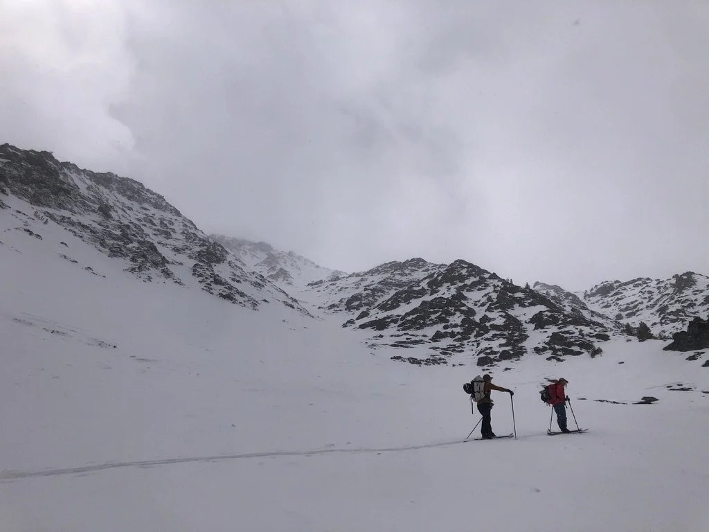 Two skiers with backpacks and poles traverse a snowy mountain landscape under a cloudy sky.