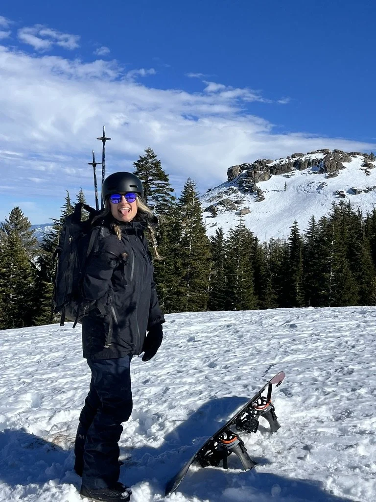 Woman dressed in black winter gear, wearing a helmet, sunglasses, and gloves, standing on snow with a snowboard in front of her, in a snowy mountainous area with evergreen trees and rocky cliffs.