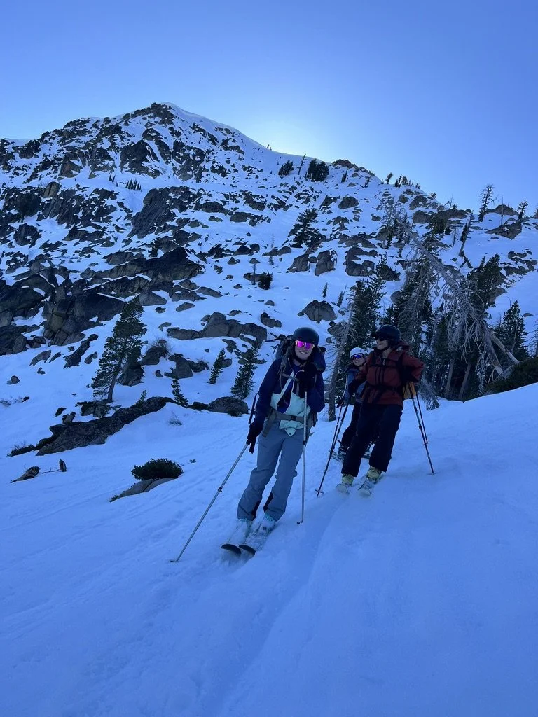 Three people snow skiing on a snowy mountain slope with a rocky, snow-covered mountain and tall trees in the background.