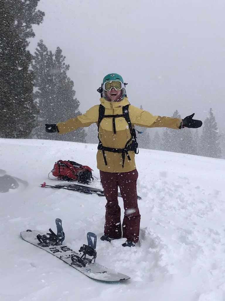Person standing in snow with arms outstretched, wearing yellow jacket, maroon pants, helmet, goggles, gloves, and ski gear, with skis and a red backpack on the ground nearby, in a snowy forested area.
