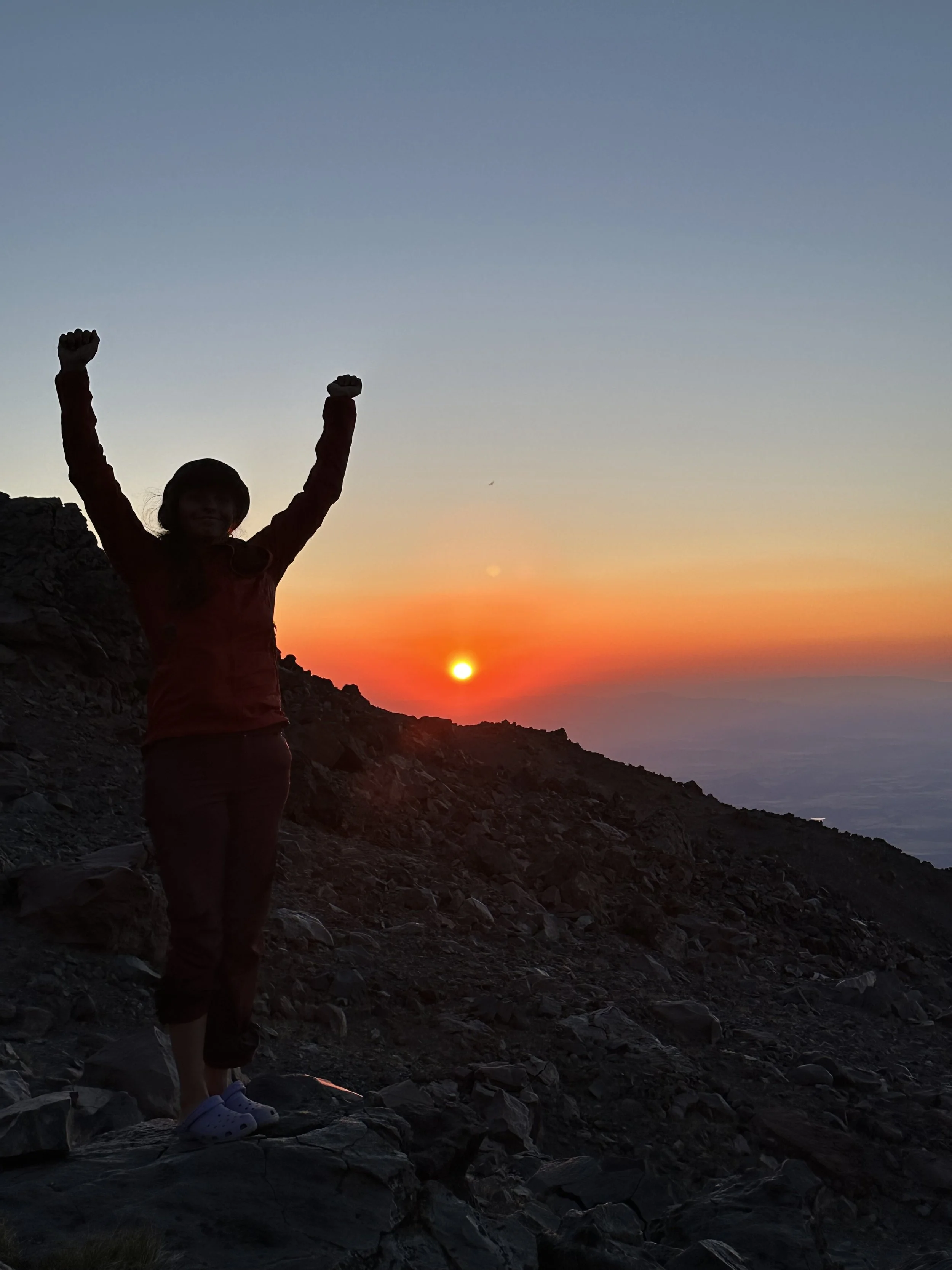 A person standing on rocky terrain with arms raised in celebration during sunset, silhouette against a colorful sky with the sun near the horizon.