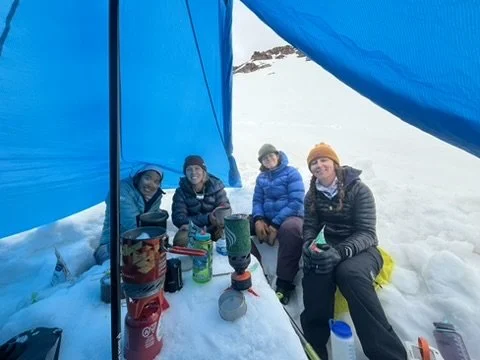 Four people inside a snow shelter with camping gear, sitting on snow.