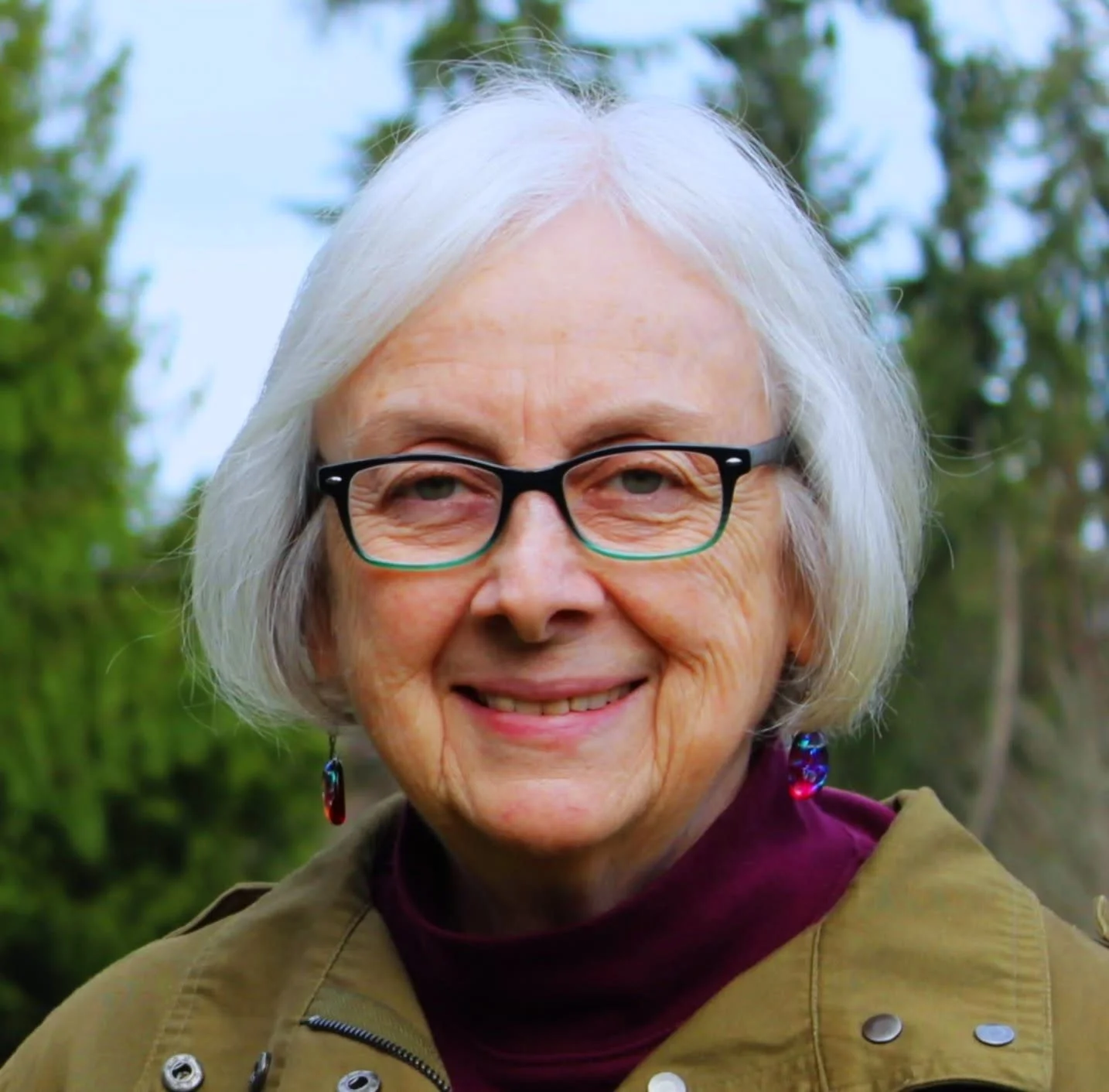 A smiling elderly woman with white hair and glasses outdoors with trees in the background.