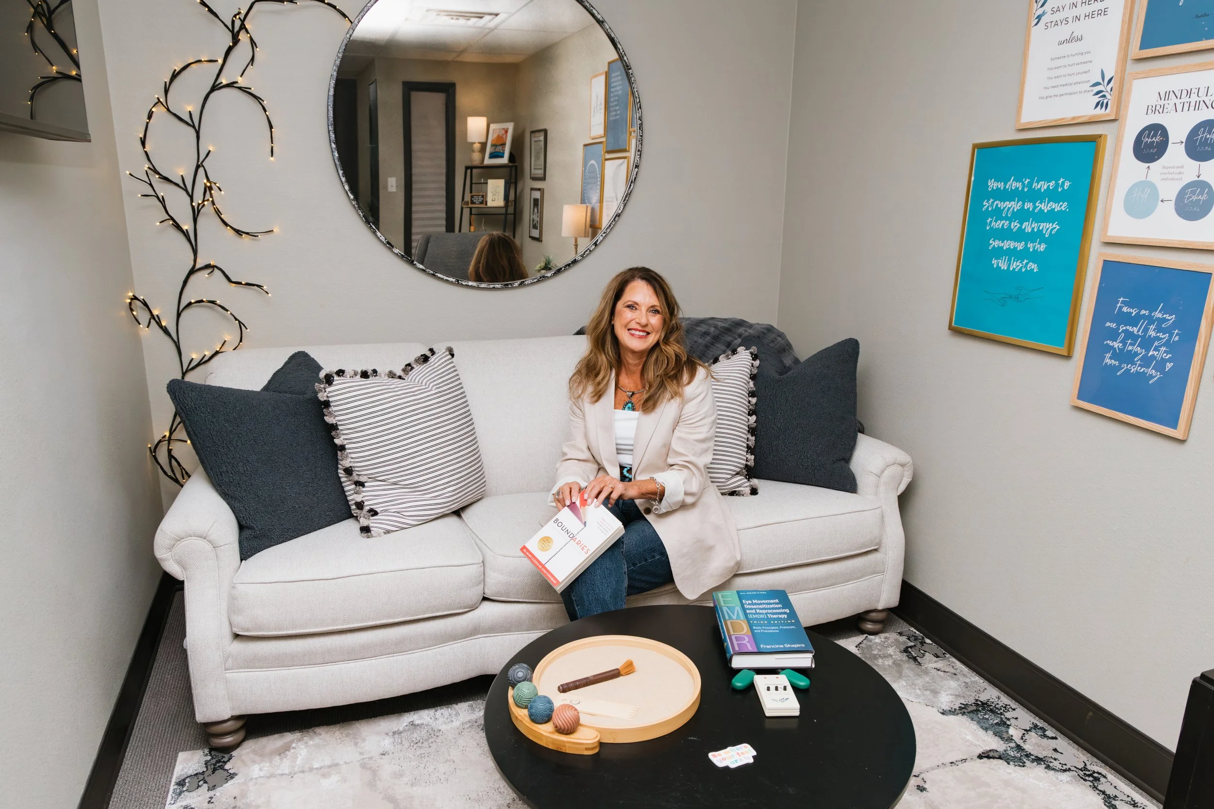 A woman sitting on a white couch holding a book, in a room with framed artwork, a mirror, and decorative string lights on the wall. There are pillows on the couch, a black coffee table with books and objects, and a patterned rug on the floor.