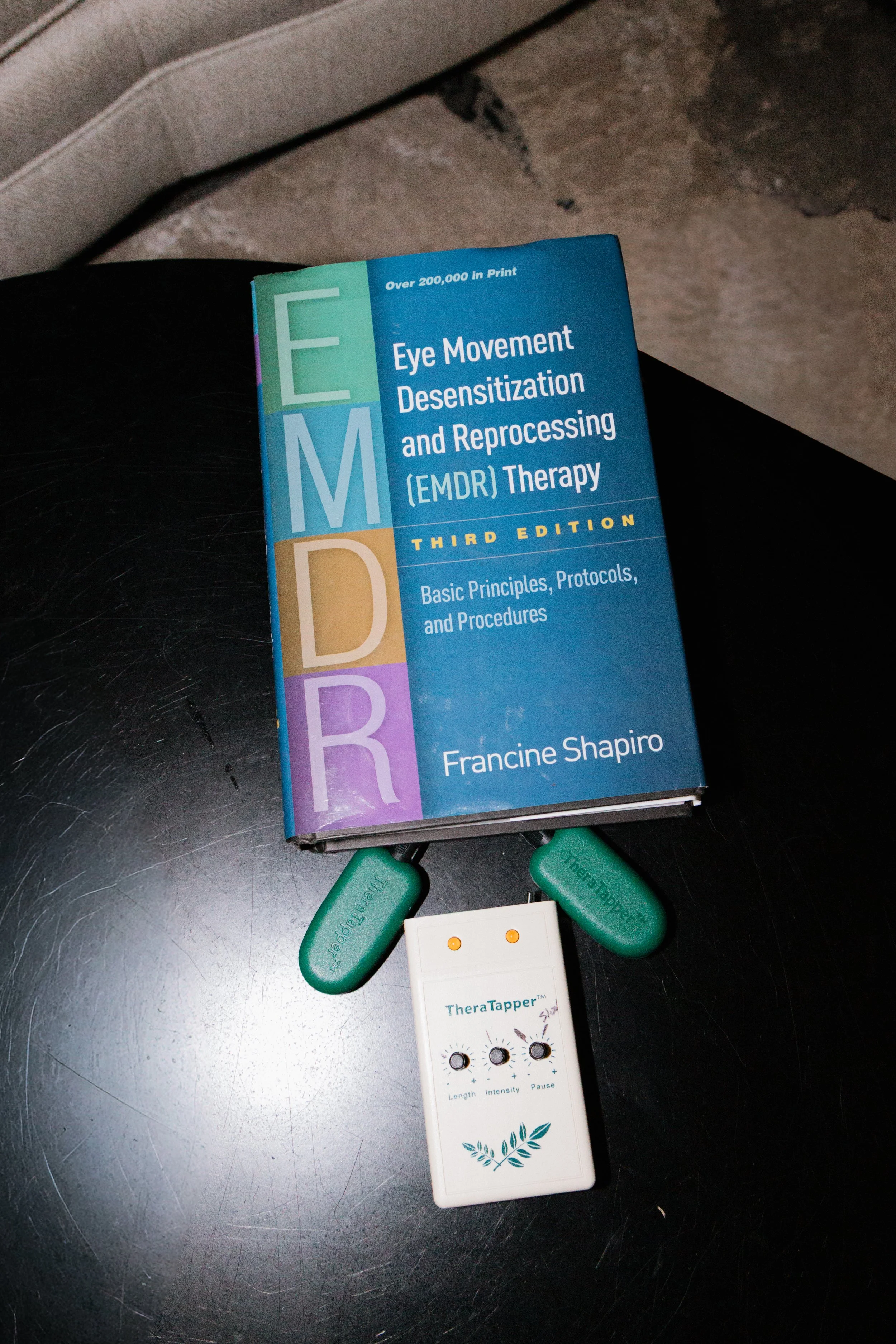 A book titled 'Eye Movement Desensitization and Reprocessing (EMDR) Therapy' placed on a dark surface, with some green sensory rings, an electric device labeled 'TheraTapper,' and a white electronic gadget with control knobs below it.