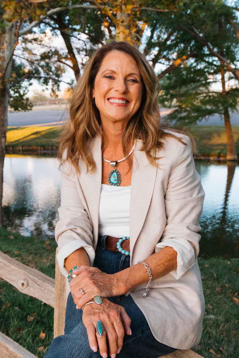A smiling woman with wavy brown hair, wearing a beige blazer, white top, and blue jewelry, sitting outdoors next to a lake with trees in the background.