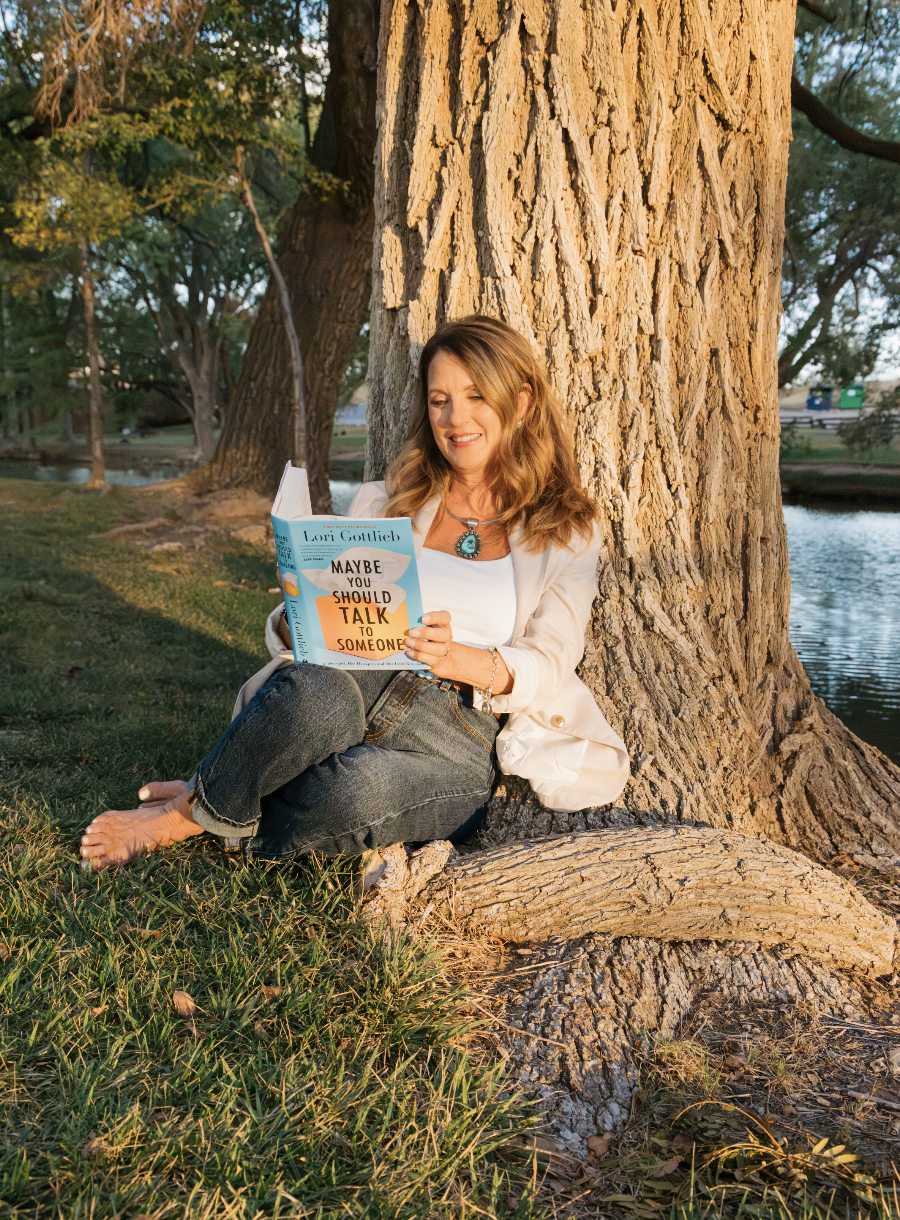 A woman with light brown hair, wearing a white jacket, white top, and blue jewelry, sitting on grass by a large tree, reading a book titled 'Maybe You Should Talk to Someone' by Lori Gottlieb, near a river at sunset.