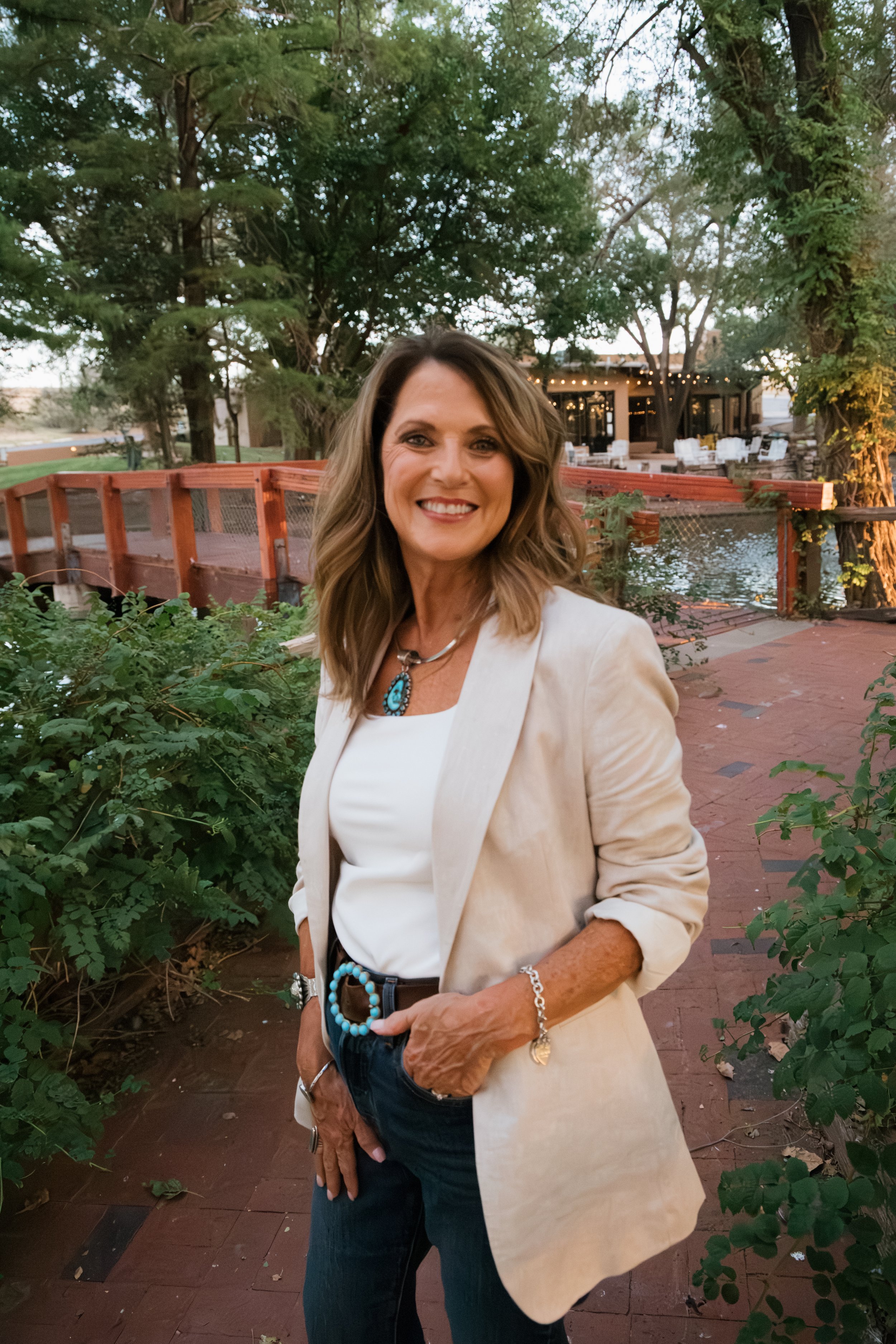 A woman with brown wavy hair, wearing a white blazer and blouse, standing outdoors at dusk near a wooden footbridge over water, with trees and a building with string lights in the background.