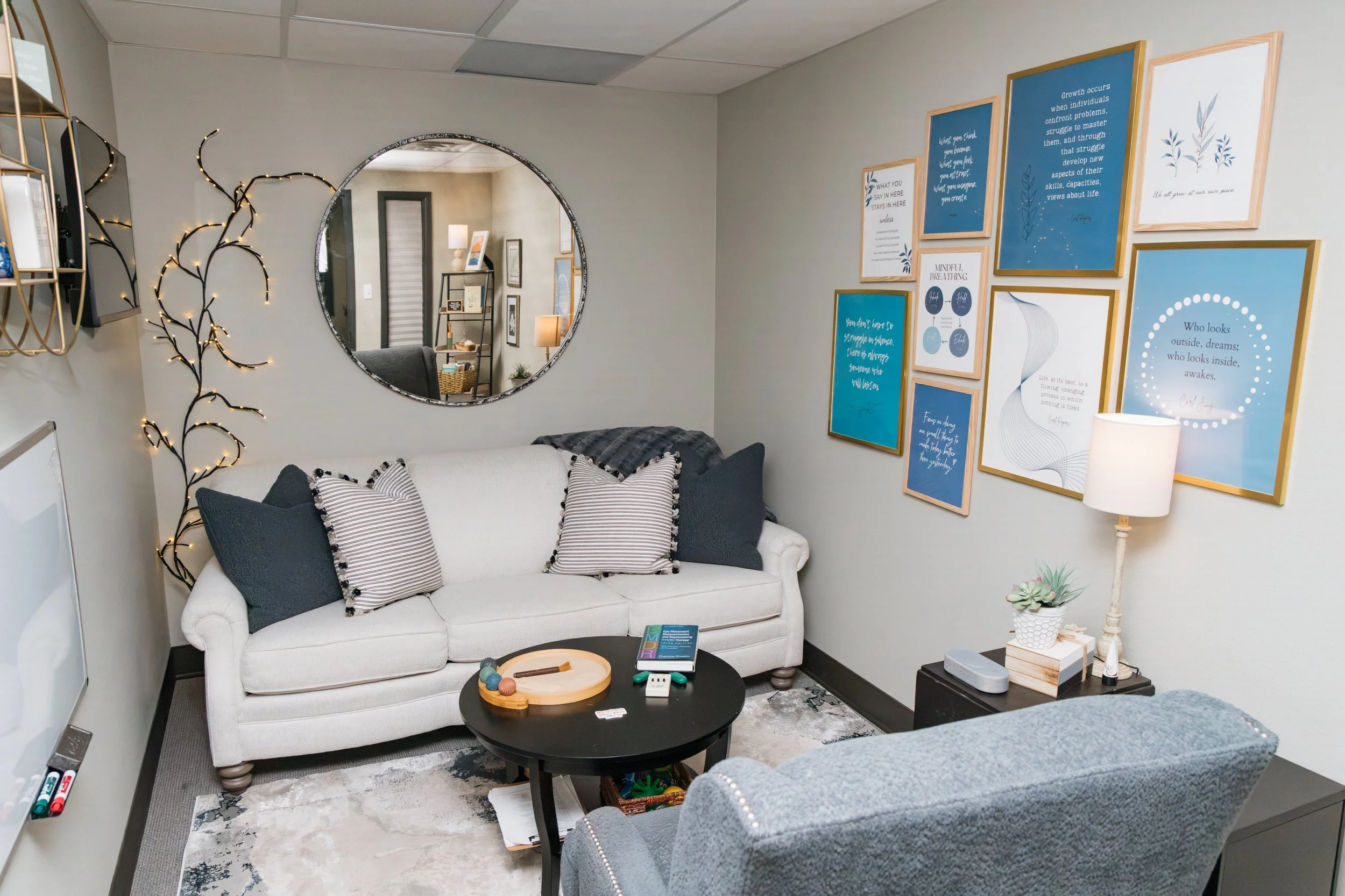 A cozy living room with a white sofa decorated with striped and solid dark cushions, a black coffee table with a game and a tray, a wall adorned with framed blue and white inspirational posters, a tall white lamp on a small side table, and a decorative branch with lights. A round mirror reflects part of the room, and a small plant is on a table near the wall.