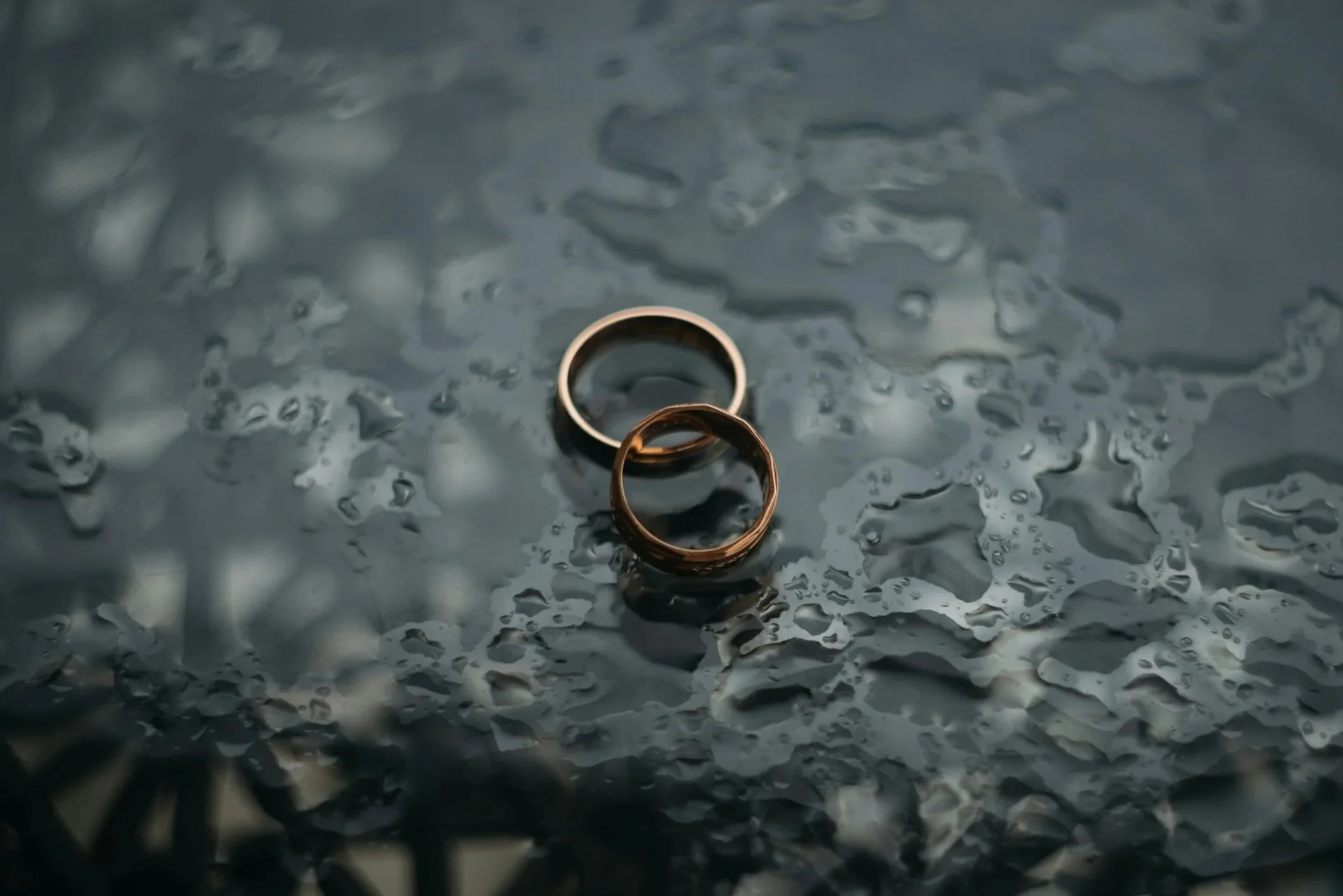 Close-up of two gold wedding rings lying on a wet, water-dappled surface.