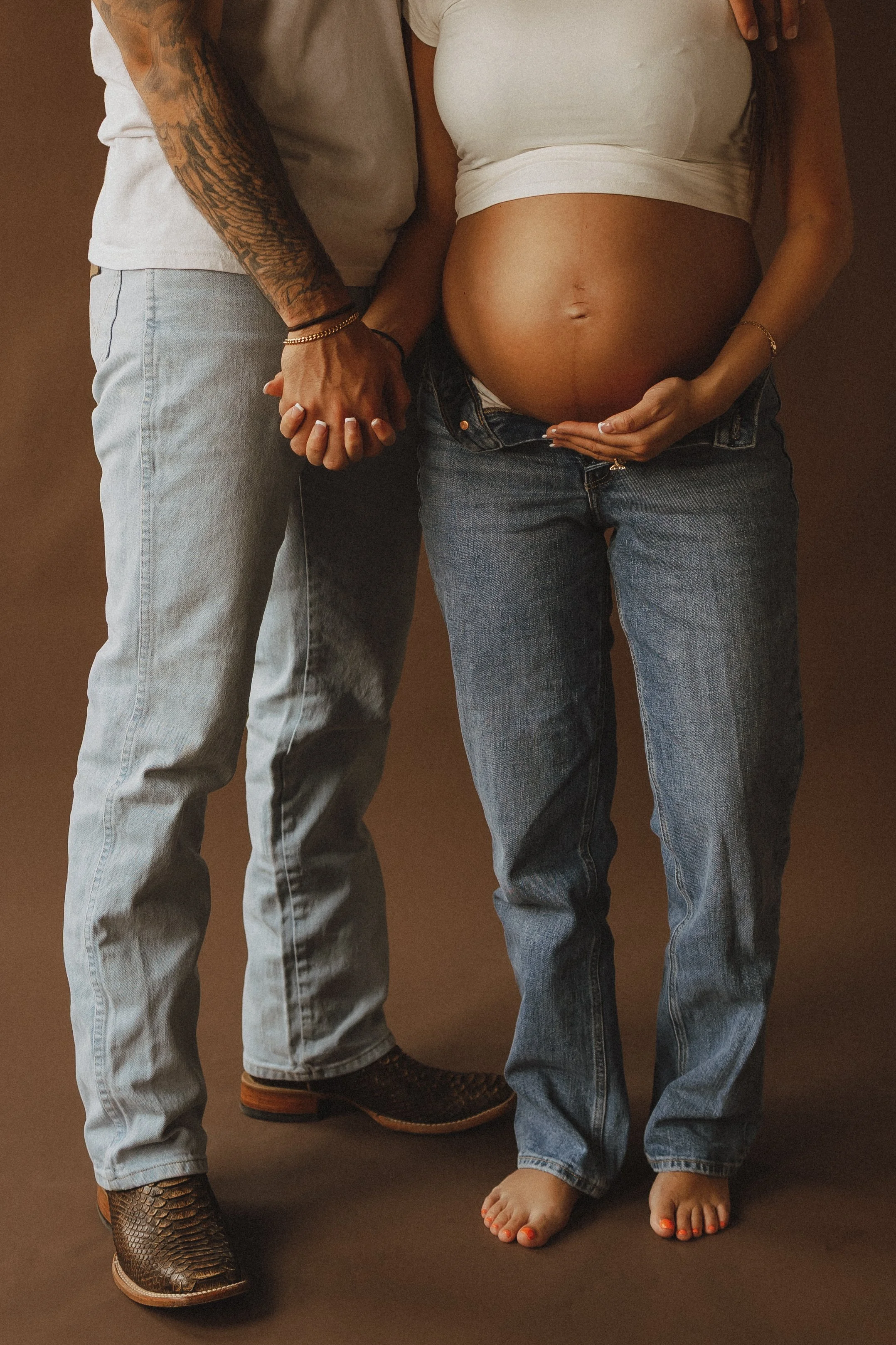 Close-up of a man and a pregnant woman holding hands, with the woman's belly in focus. They are standing against a plain brown background, and both are dressed casually in jeans.
