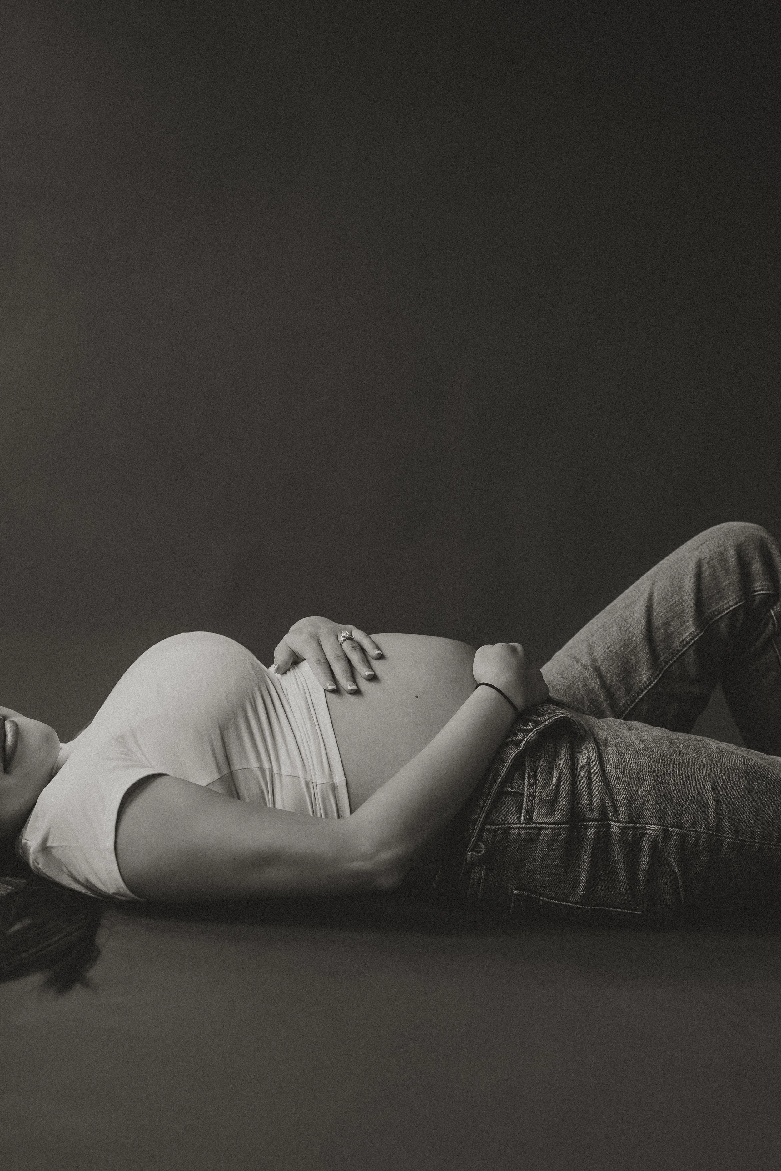 A woman lying on her back on the floor, holding her pregnant belly with both hands, wearing a white t-shirt and jeans.