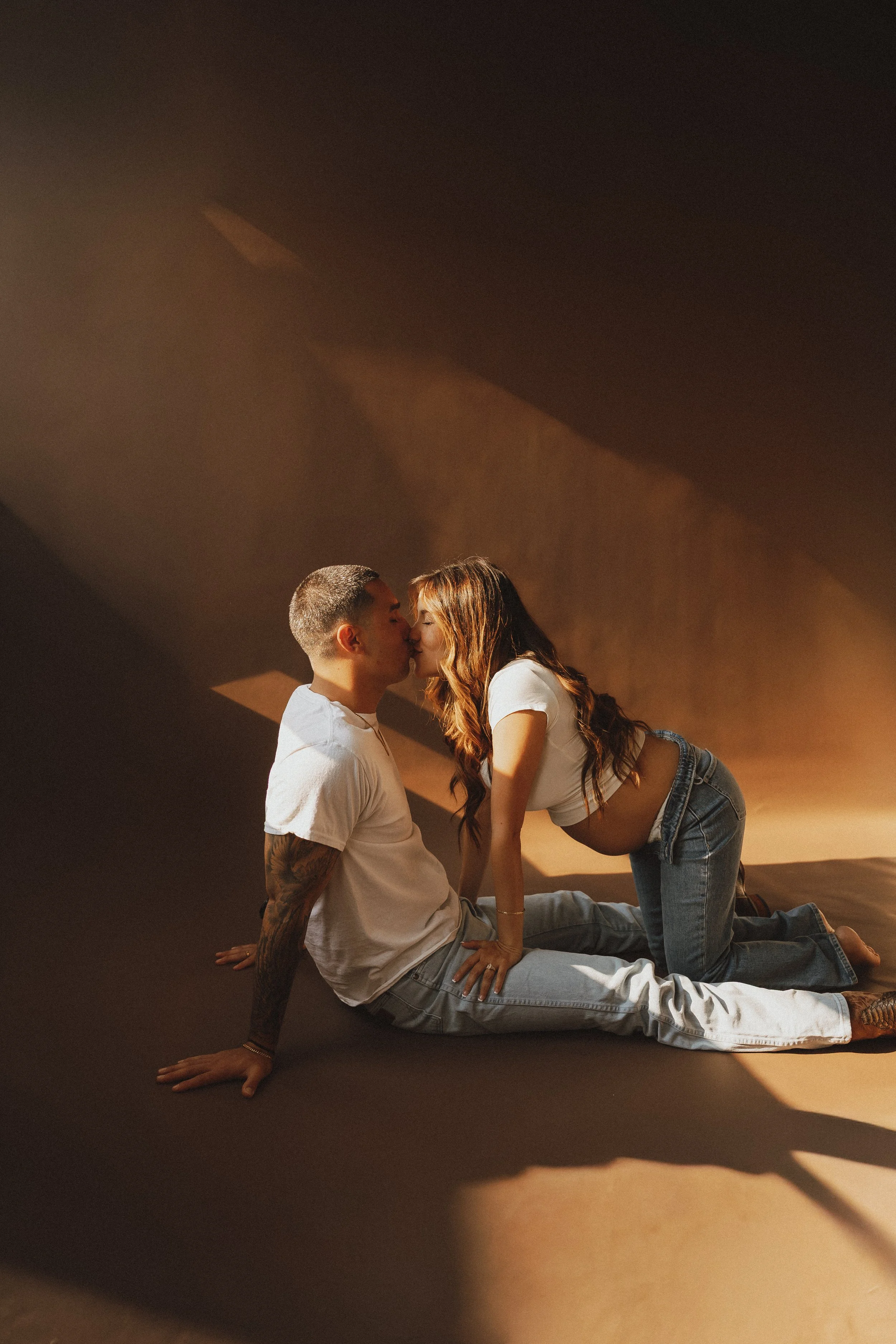 A couple is positioned intimately close, with the woman leaning toward the man for a kiss. They are indoors with warm lighting, and the scene appears romantic and affectionate.