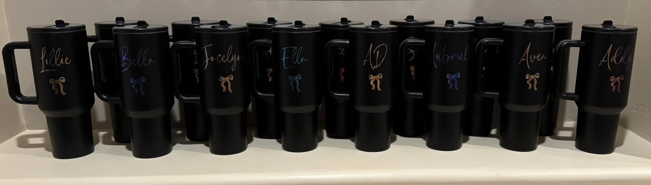 Black mugs with colorful names and bows, arranged on a shelf.
