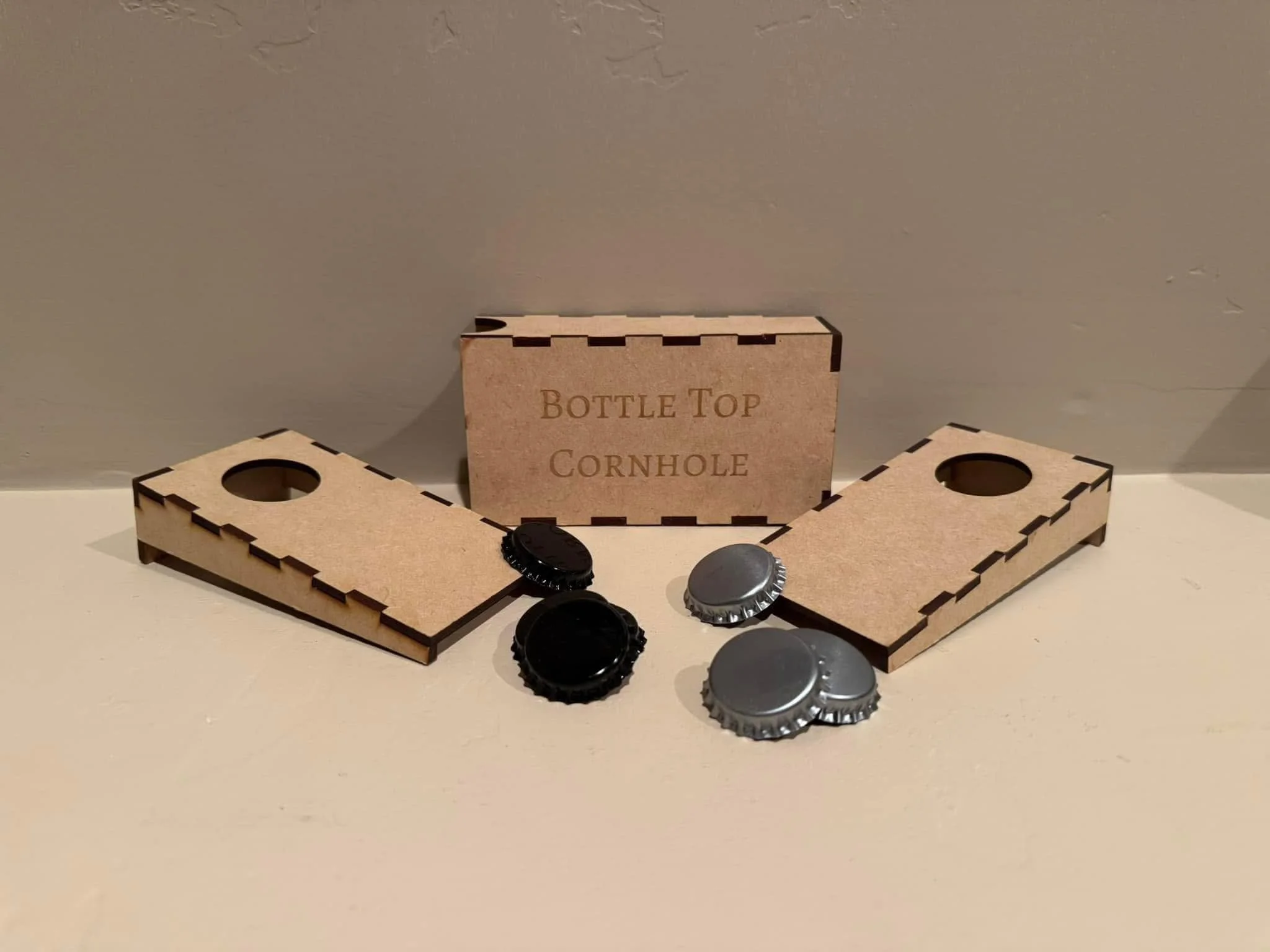 A cornhole game set with a wooden board and a box labeled 'Bottle Top Cornhole,' along with several bottle caps placed on and around the boards on a white surface against a plain wall.
