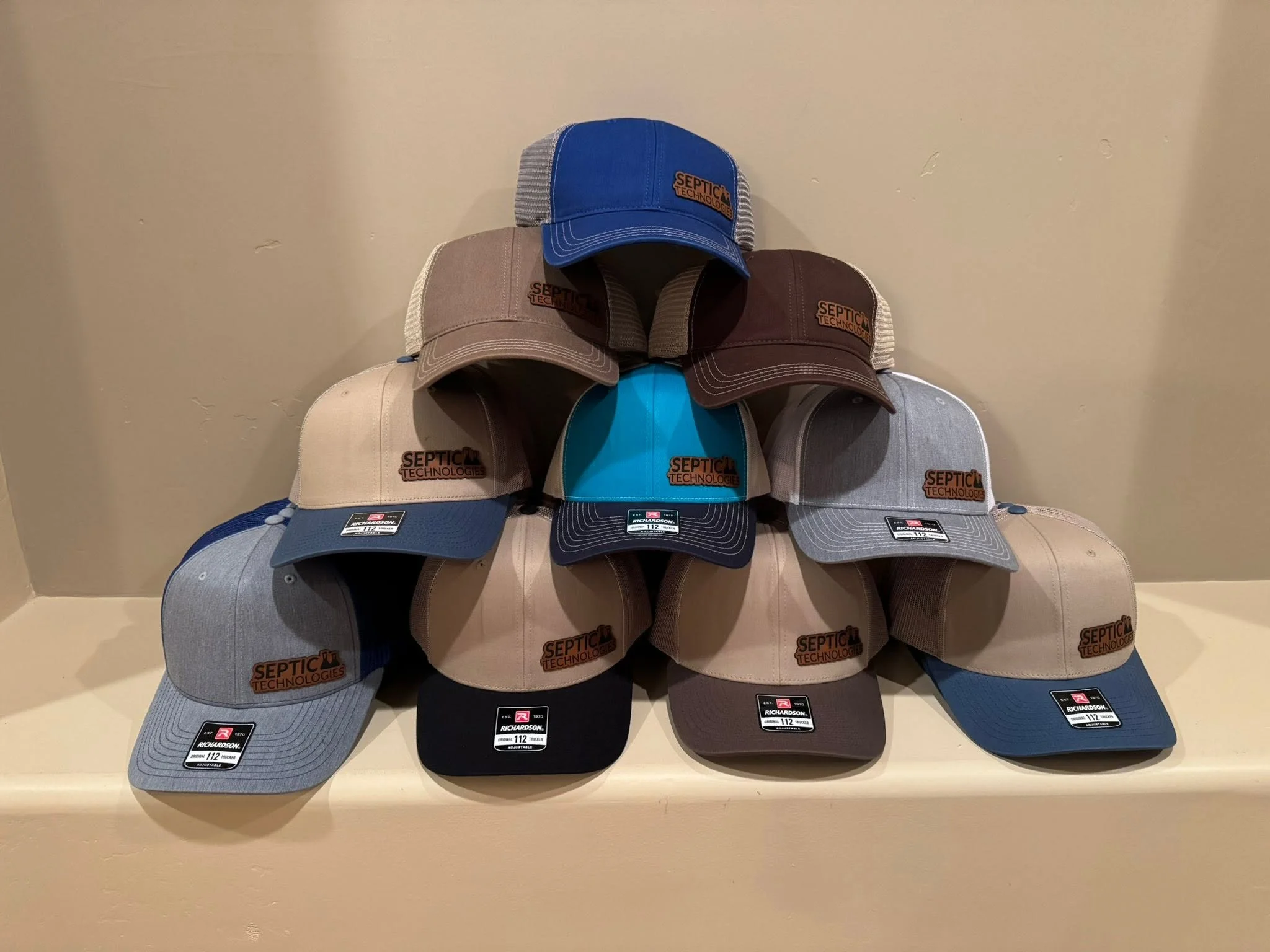 Display of multiple trucker hats with the septic technologies logo, arranged in two rows on a beige surface against a plain wall.