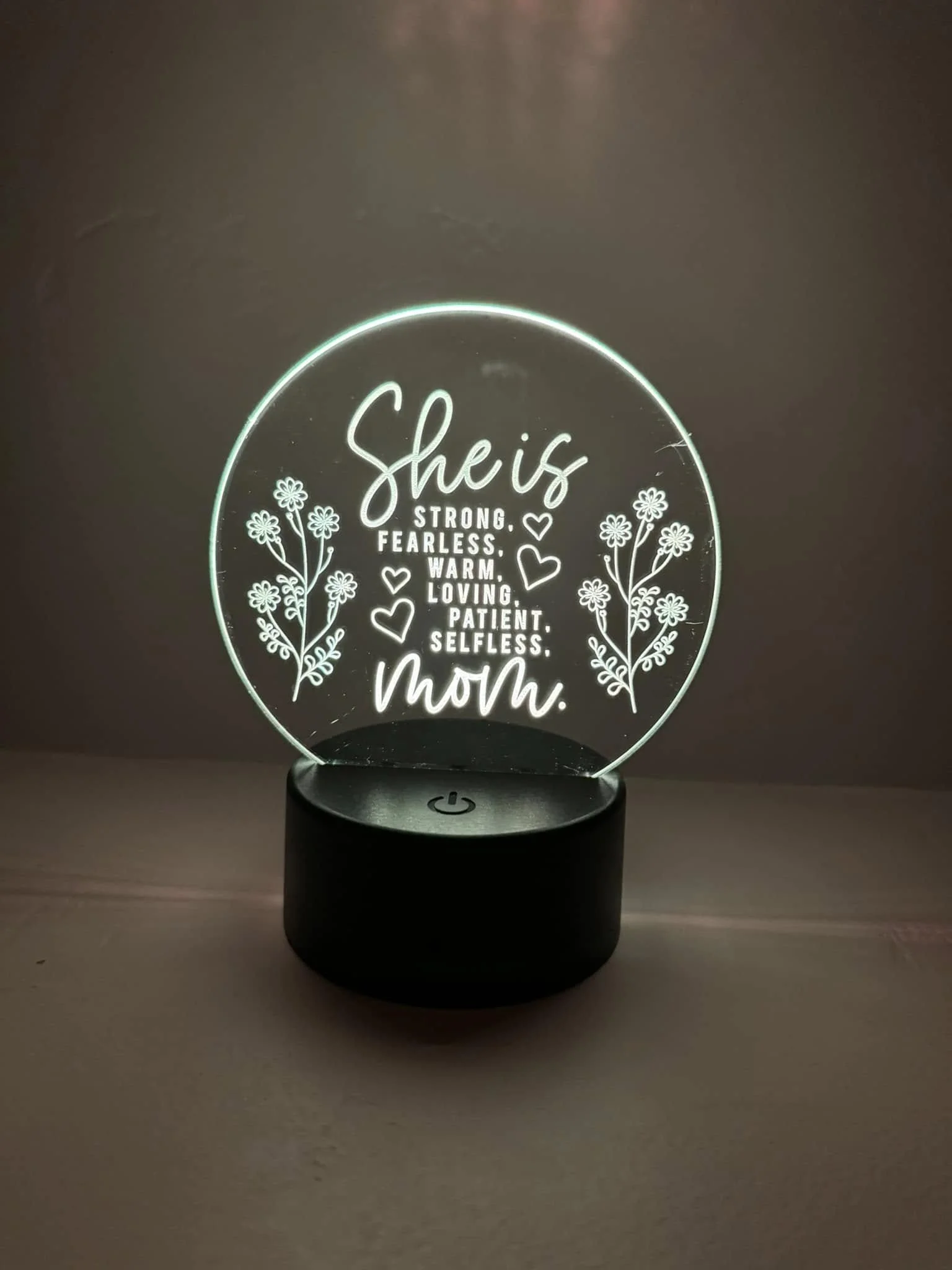 A decorative lamp with a circular glass panel inscribed with inspirational words like "She is strong, fearless, warm, loving, patient, selfless" and adorned with floral designs, placed on a dark base with a power button.