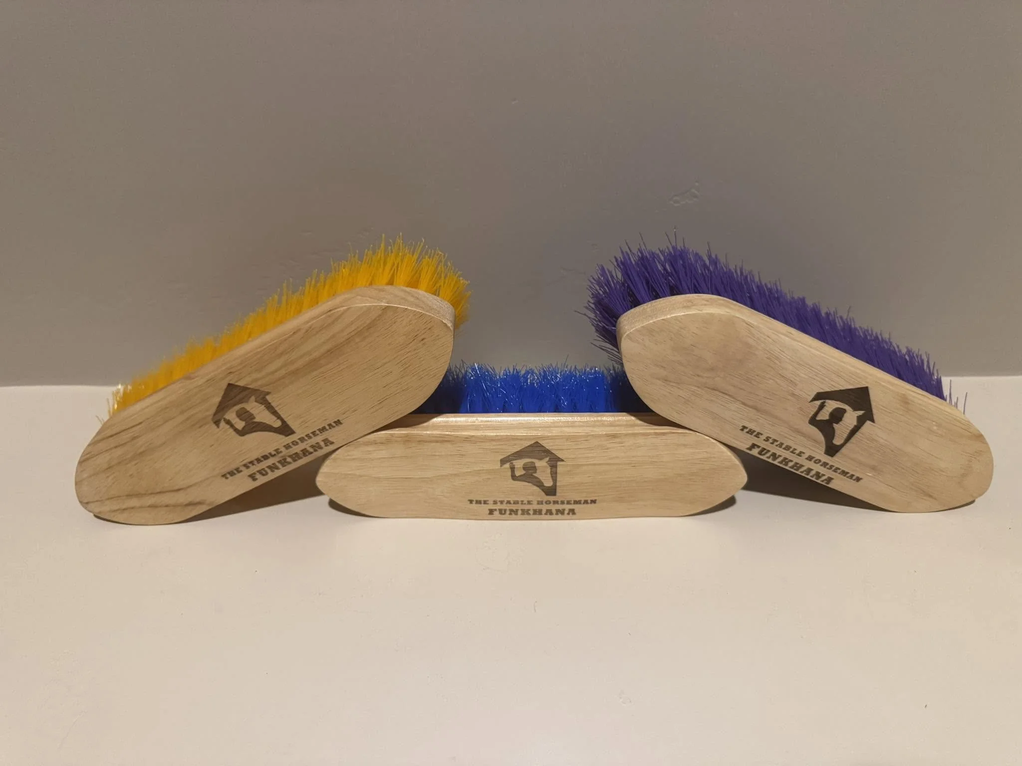 Three cleaning brushes with wooden handles and colorful bristles, yellow on the left, purple on the right, and blue in the middle, on a white surface against a gray wall, with logo and text on handles.