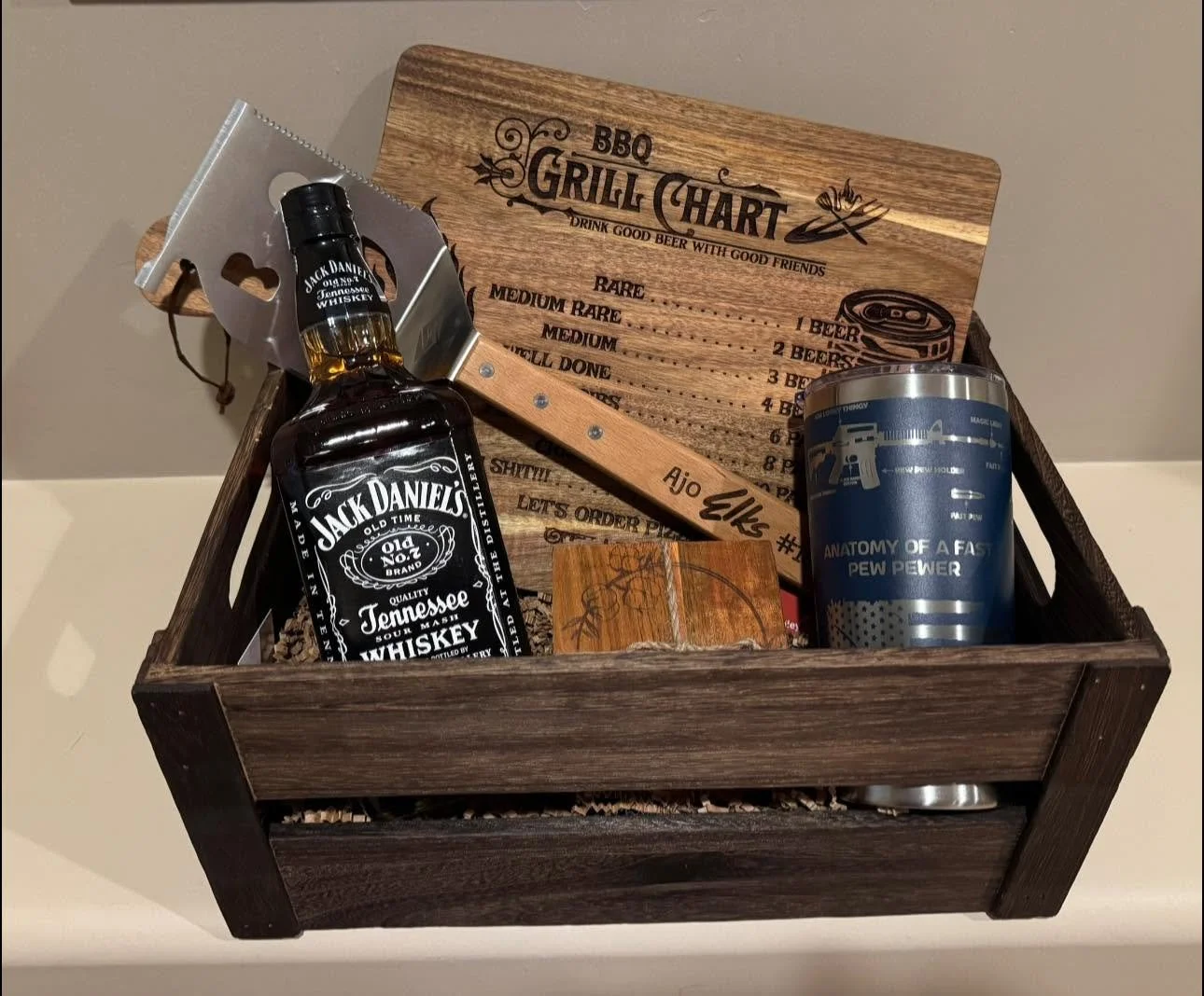 A wooden gift basket containing a bottle of Jack Daniel's whiskey, a beverage tumbler with a design, a small decorative box, and a wooden sign with a barbecue theme and a handwritten name.