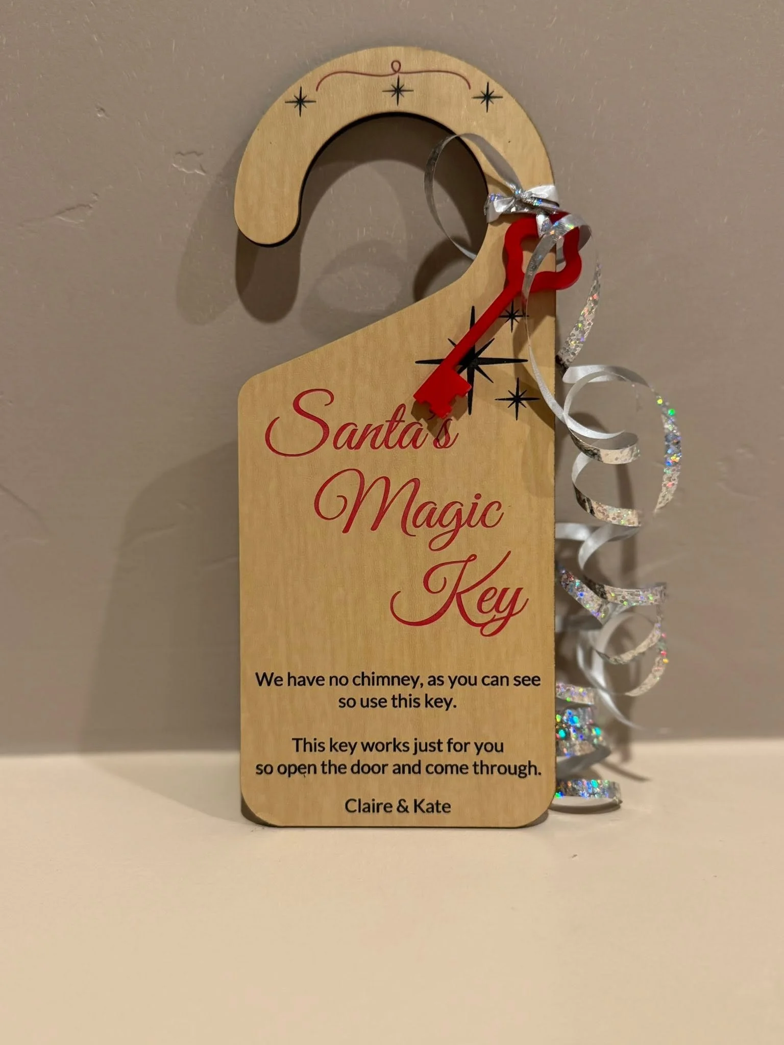 A wooden key-shaped sign with red and black text that reads, "Santa's Magic Key." The sign has decorative black stars and red accents, and is attached to metallic silver and red curly ribbons. Below, at the bottom of the sign, there is text that expl
