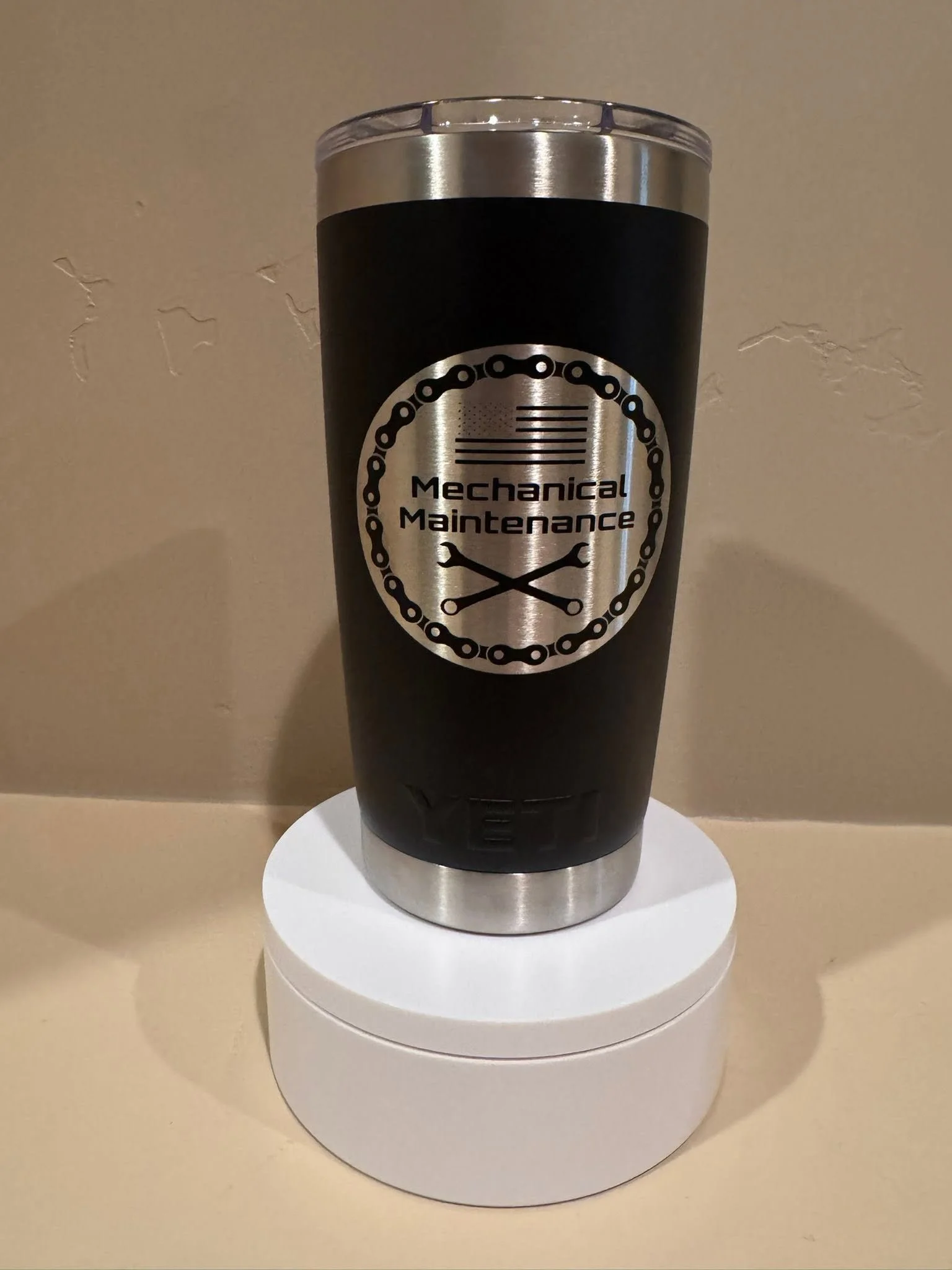 Travel mug with a black body, silver top and bottom, and a logo that reads 'Mechanical Maintenance' with a wrench and gear outline, placed on a white cylindrical stand.