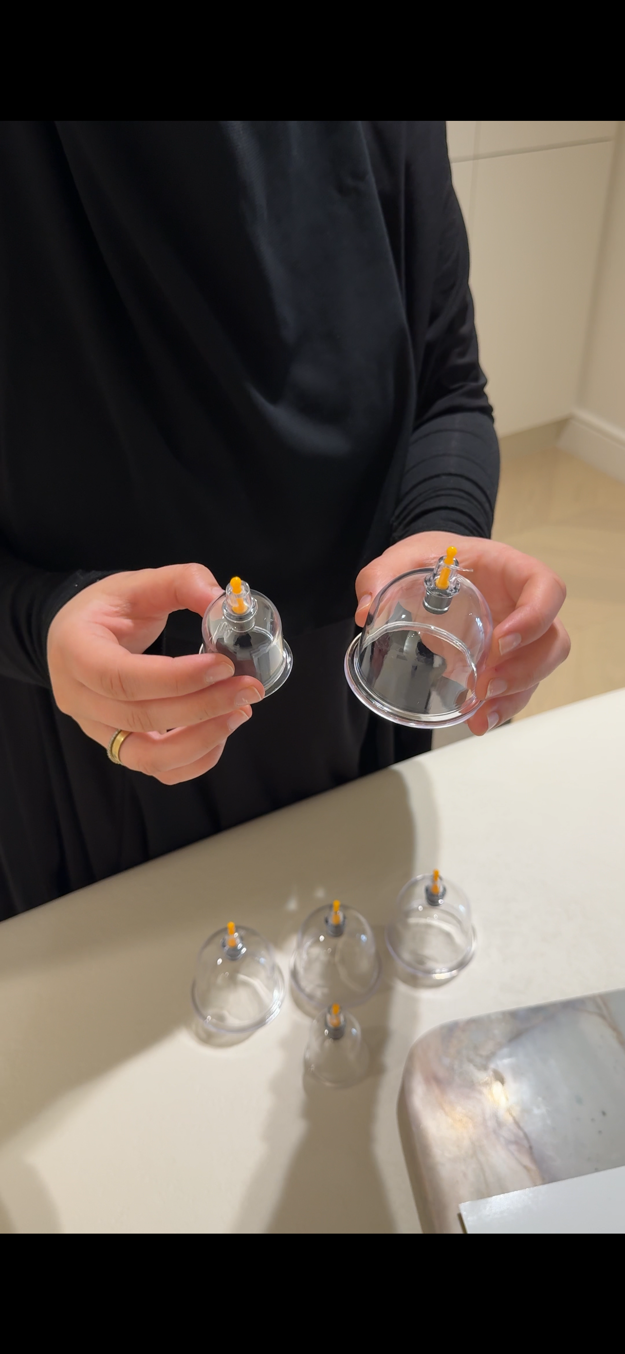 Person holding small inverted clear plastic cups with syringe-like applicators on top, possibly for skincare or cosmetic treatment, on a kitchen countertop.