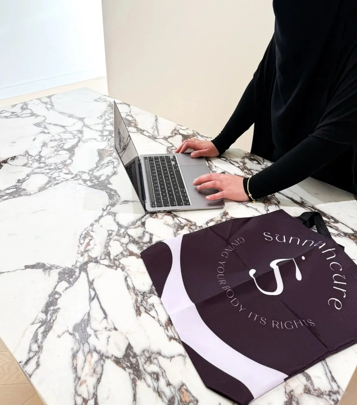 A person working on a laptop at a marble table with a purple and white tote bag that says 'Sunshine' and 'Giving your body its rights'.