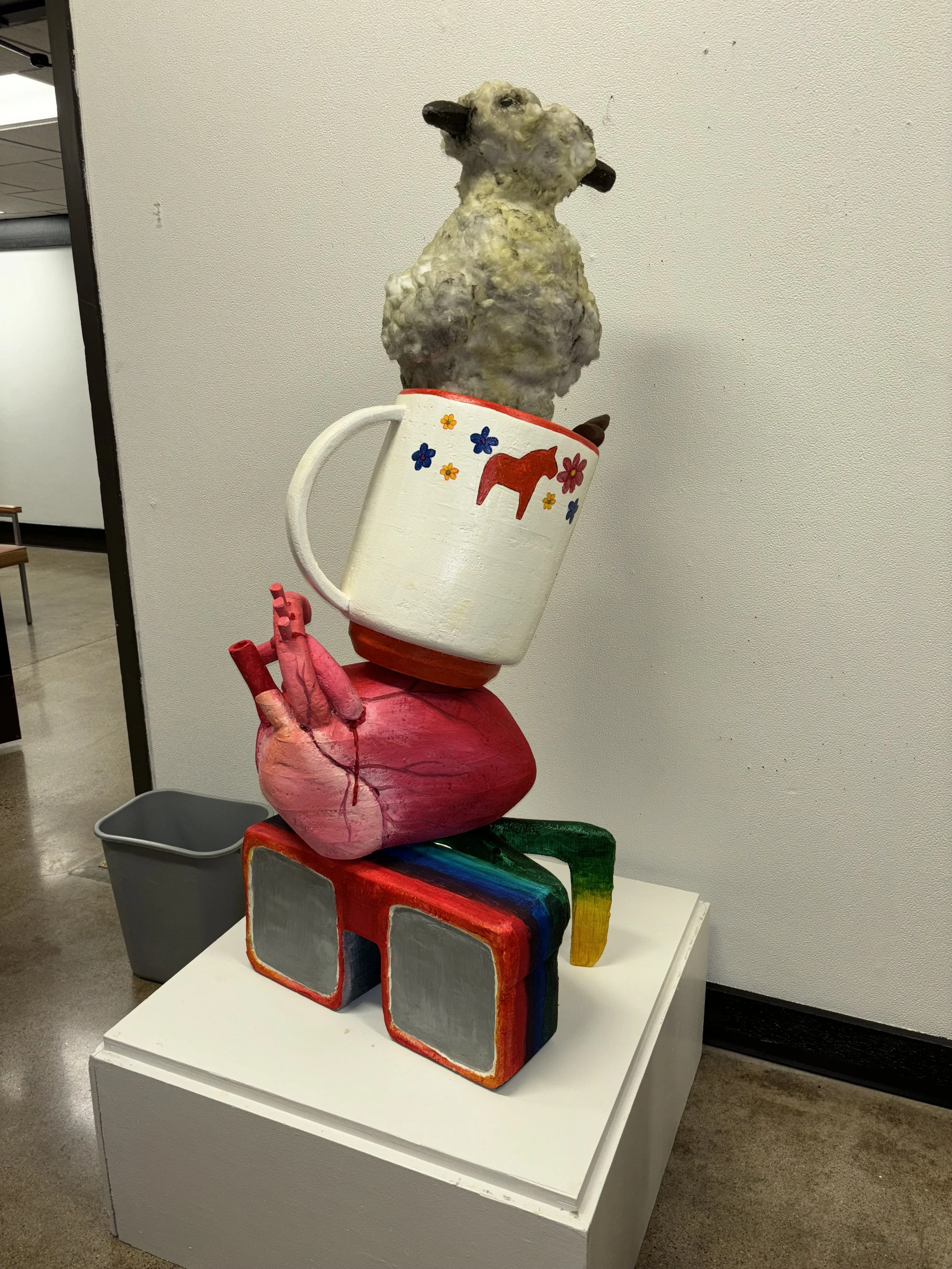 A colorful sculpture of a pair of glasses underneath a heart with a mug on top, which is filled with a sheep's head and body, and has a red horse painted on the mug. The sculpture is displayed on a white table.