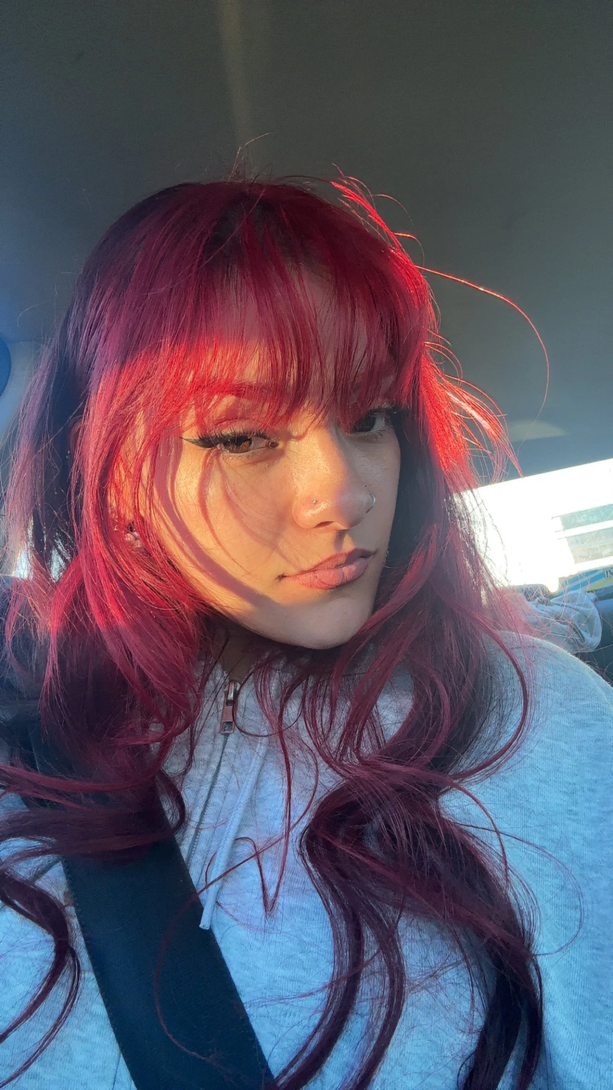 A woman with long, wavy red hair, wearing makeup and a nose ring, sitting in a car with sunlight casting shadows on her face.