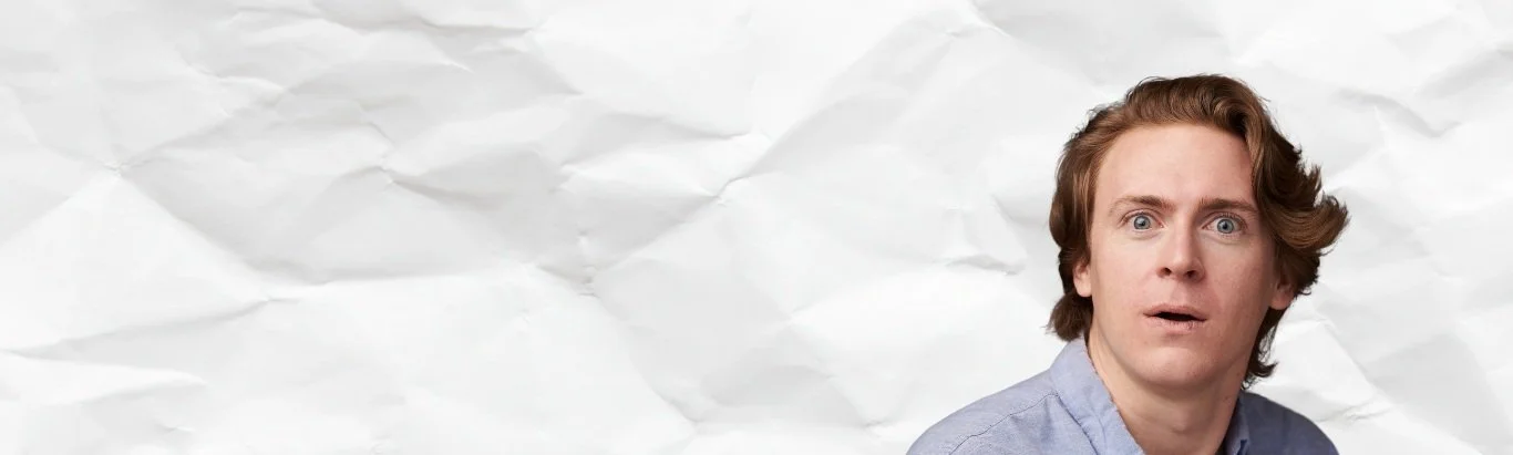 A young man with blonde hair and blue eyes looking surprised or confused, wearing a light blue shirt against a crumpled white paper background.