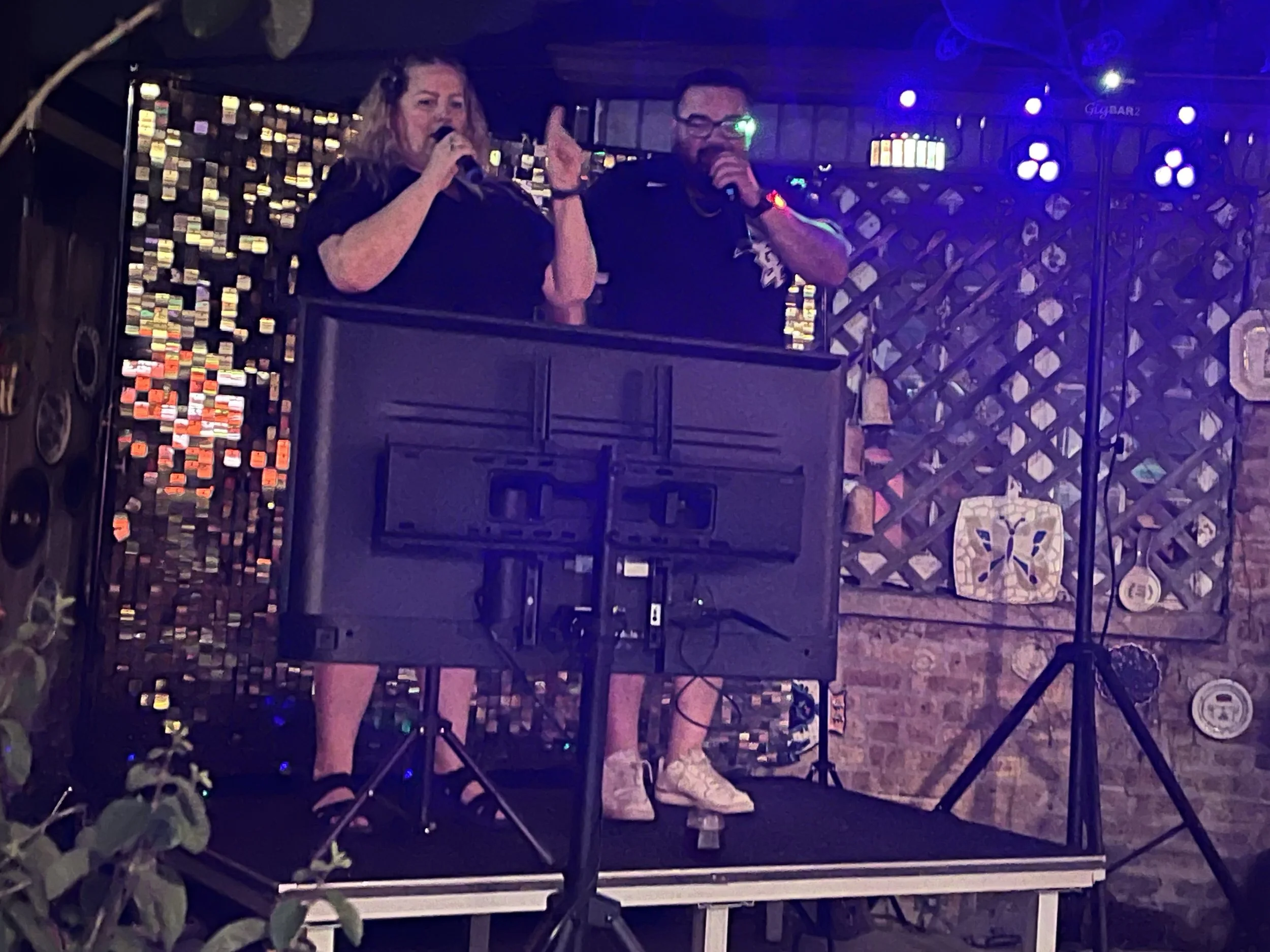 Two performers, a woman and a man, singing into microphones on a small stage in a dimly lit nightclub or bar. The woman has blonde hair, is wearing a black shirt, and the man has dark hair, glasses, and is dressed in black. Stage lights and decorative elements are visible in the background.